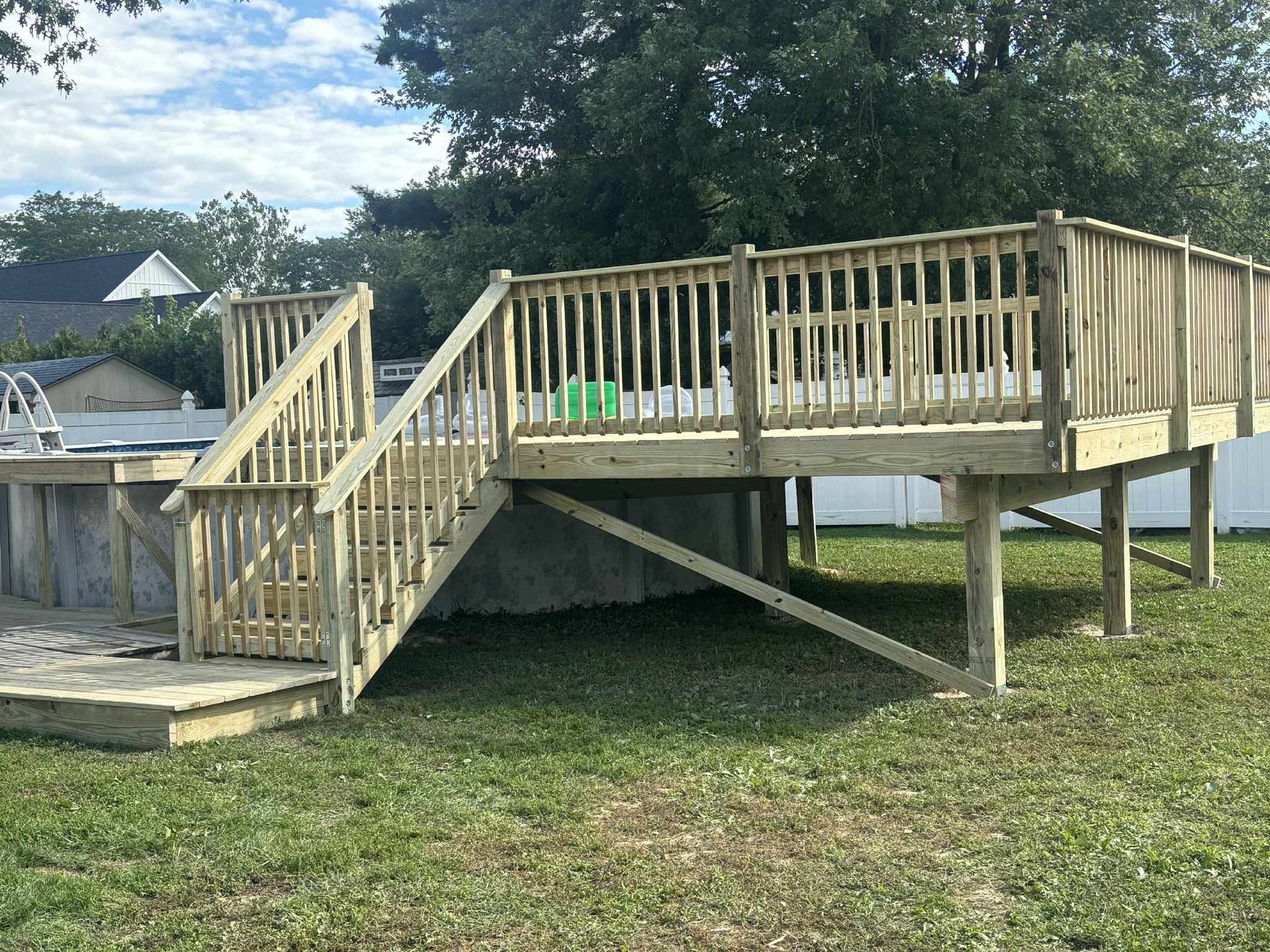 Wooden deck with stairs next to an above-ground pool, in a grassy backyard, on a sunny day.