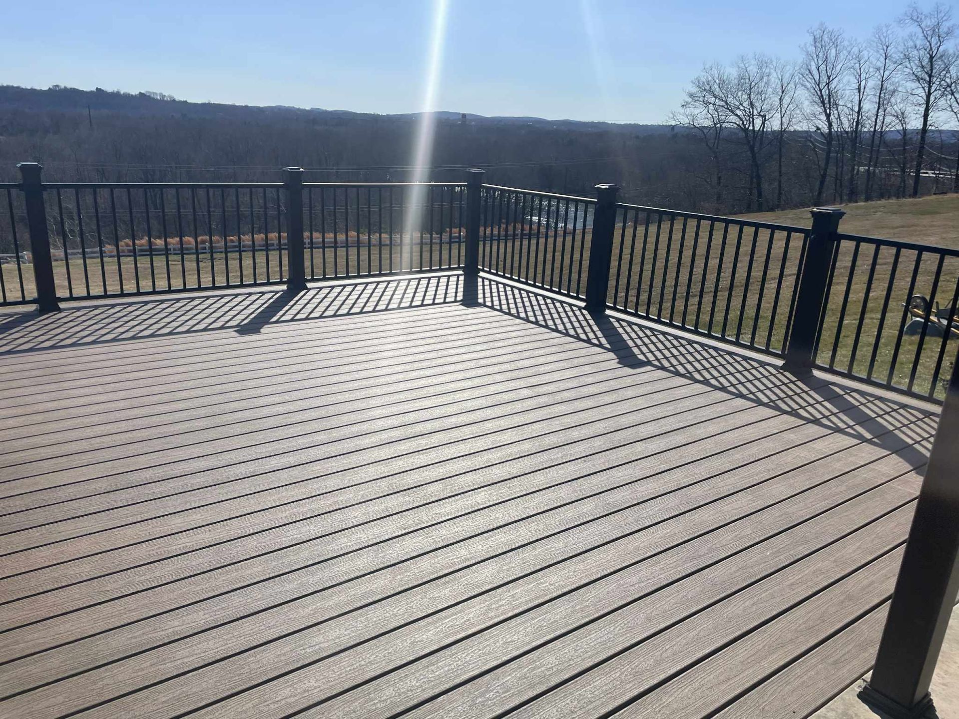 Brown composite deck with black railing overlooking a landscape with trees and a bright sky.