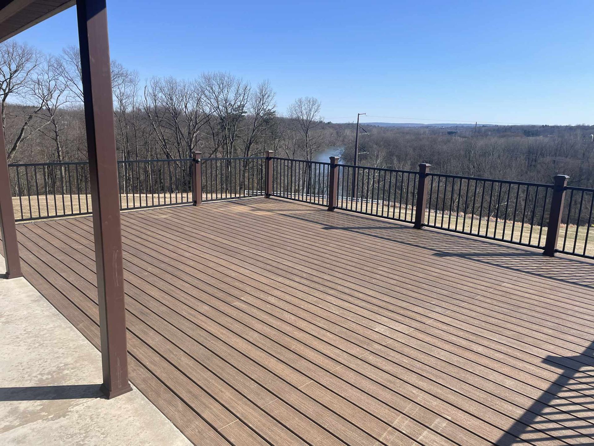 Wooden deck with dark railing overlooks a forest on a sunny day.