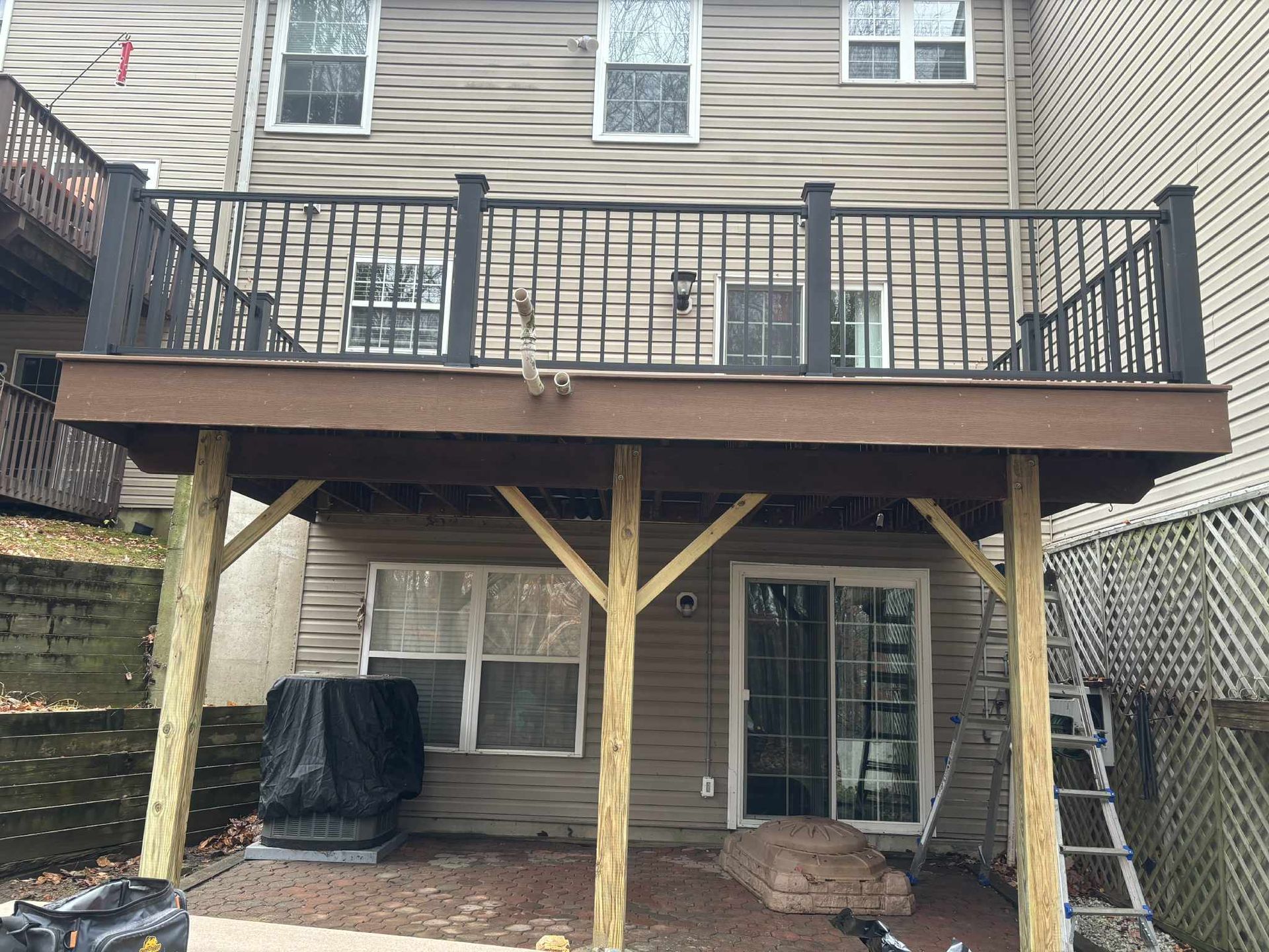 A deck with dark railings and support beams, overlooking a patio area. The home has vinyl siding.