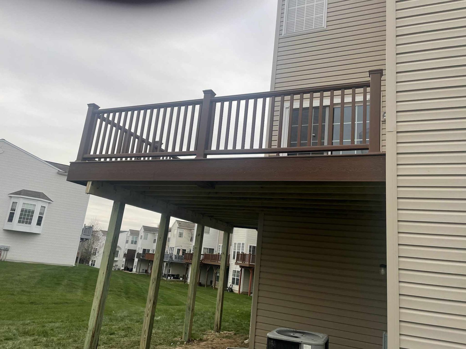 Brown deck with railing attached to a beige house, supported by wooden posts in a grassy yard.