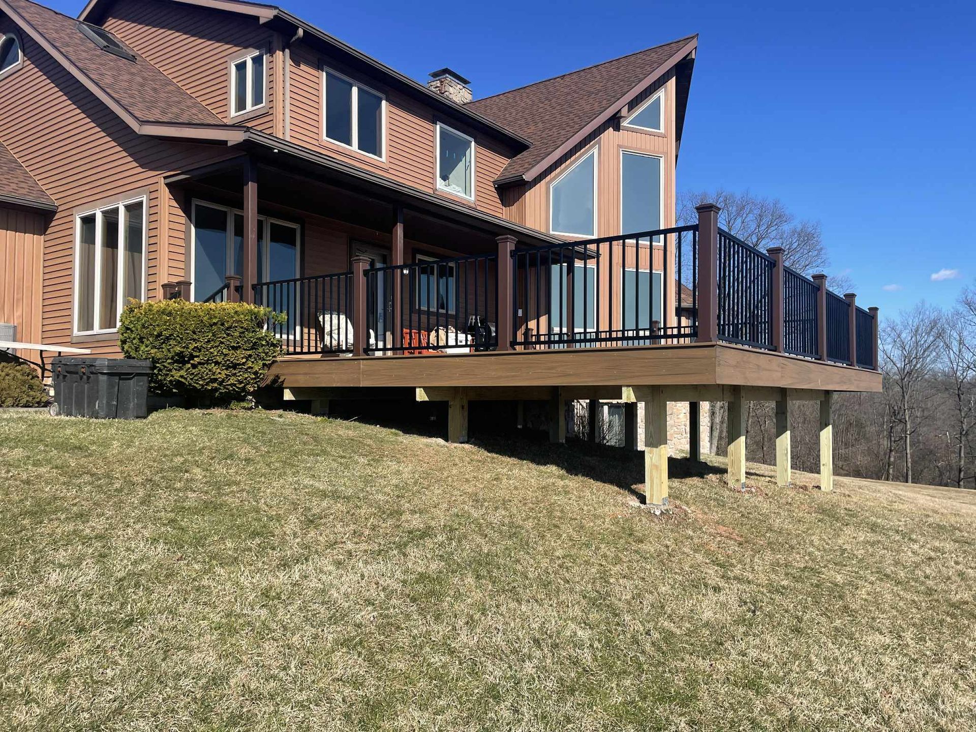 House with a brown roof and a composite deck on elevated wooden posts, overlooking a grassy hill.
