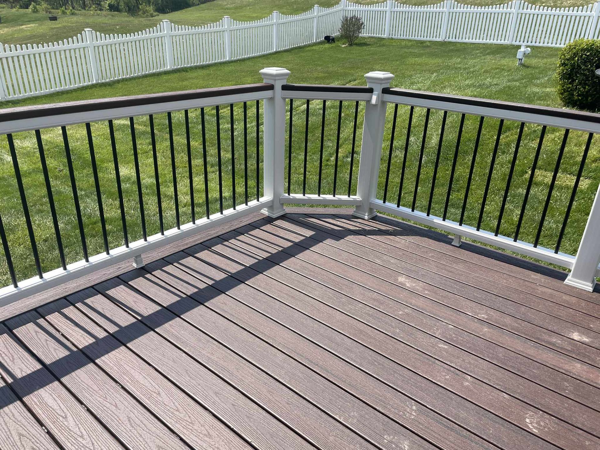 A deck with brown composite decking, white railing with black spindles, and a white picket fence in the background.