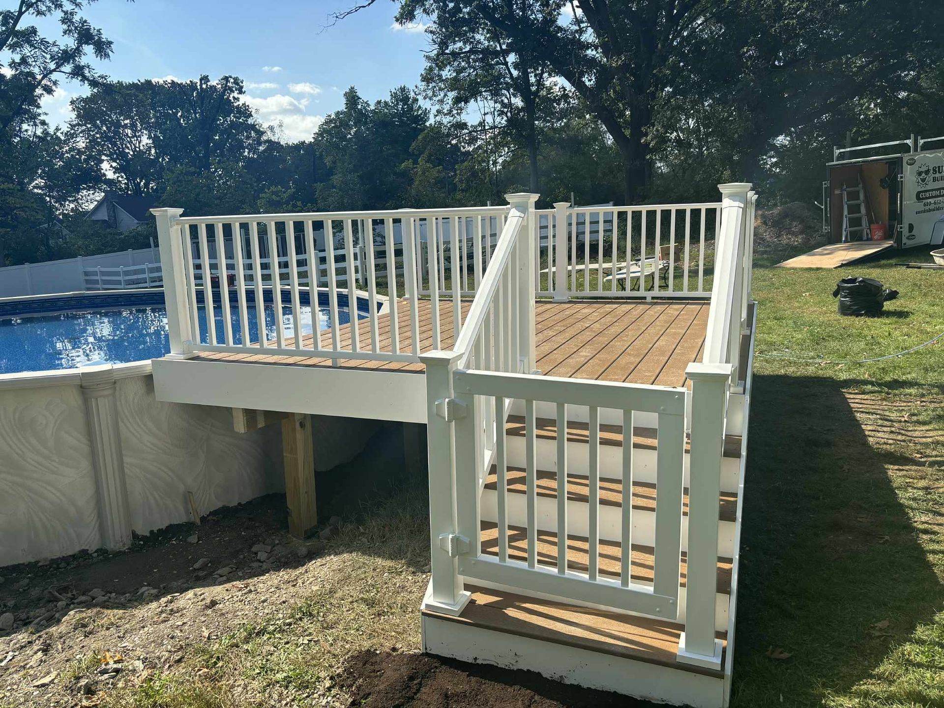 White-railed deck with stairs next to a pool in a backyard. Brown decking, green grass, and trees in background.