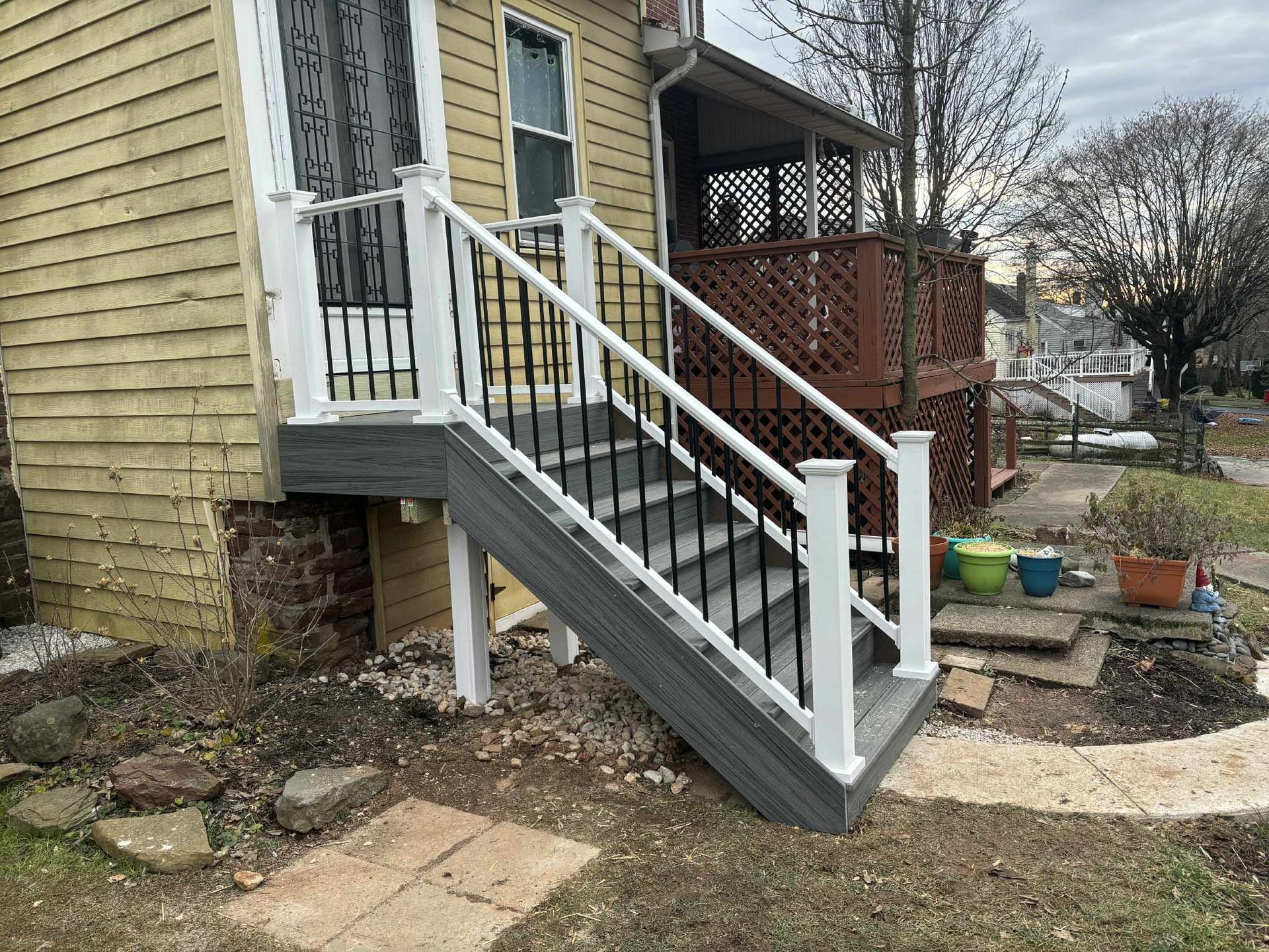 Wooden deck with white railings and dark gray steps leading up to a yellow house with a porch.