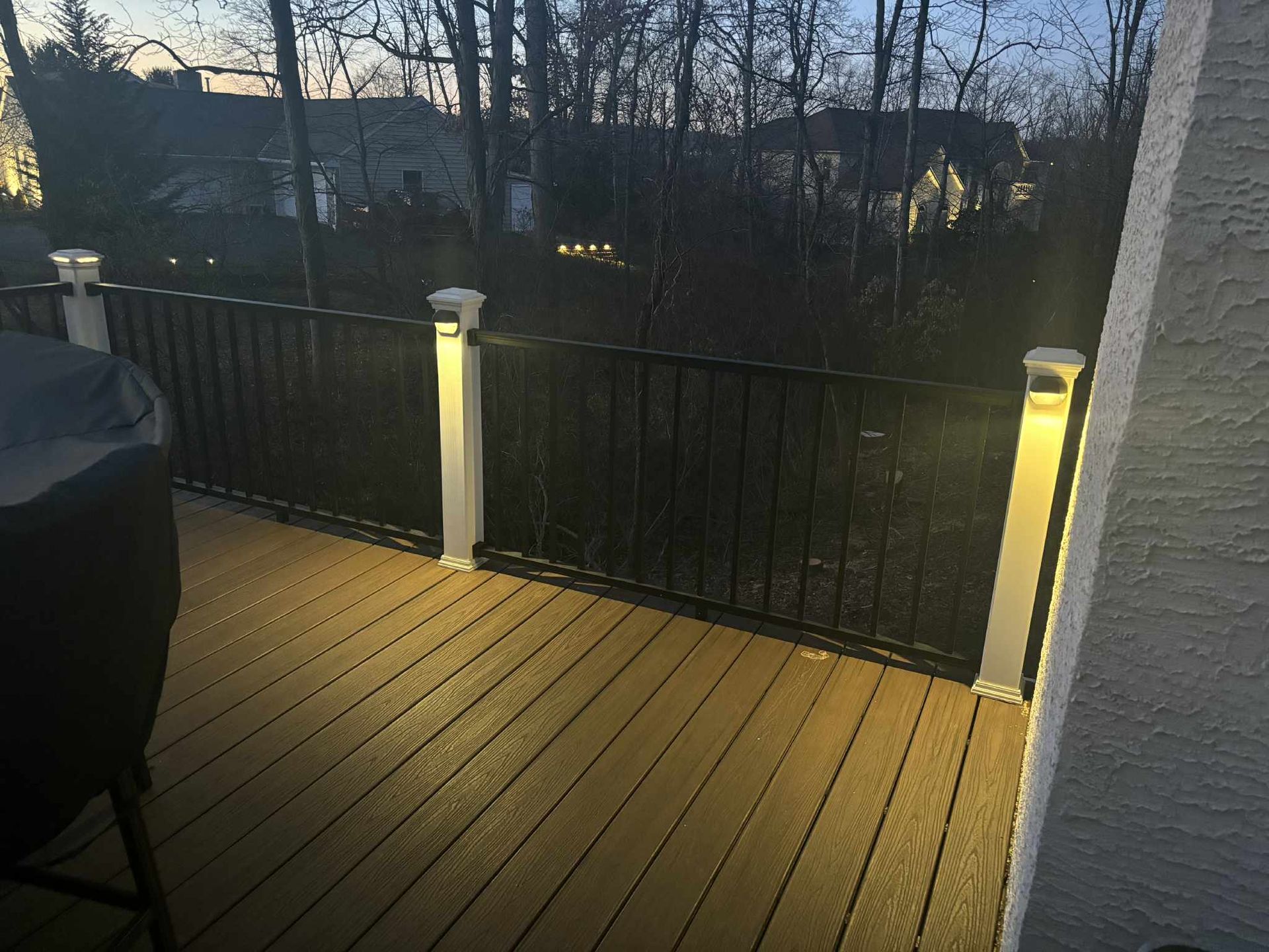 Lit deck at dusk with black railing, white posts with lights, and trees in the background.
