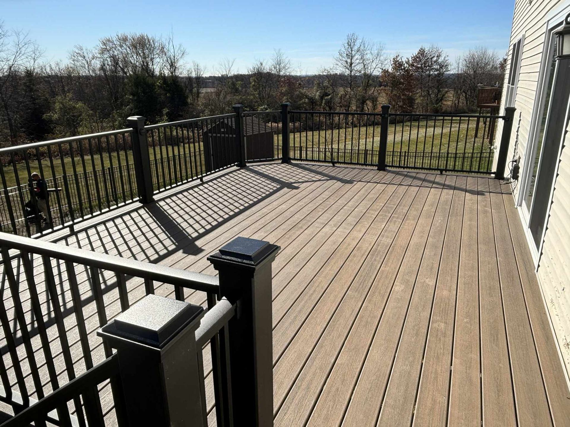 Wooden deck with black railings overlooking a grassy field under a blue sky.