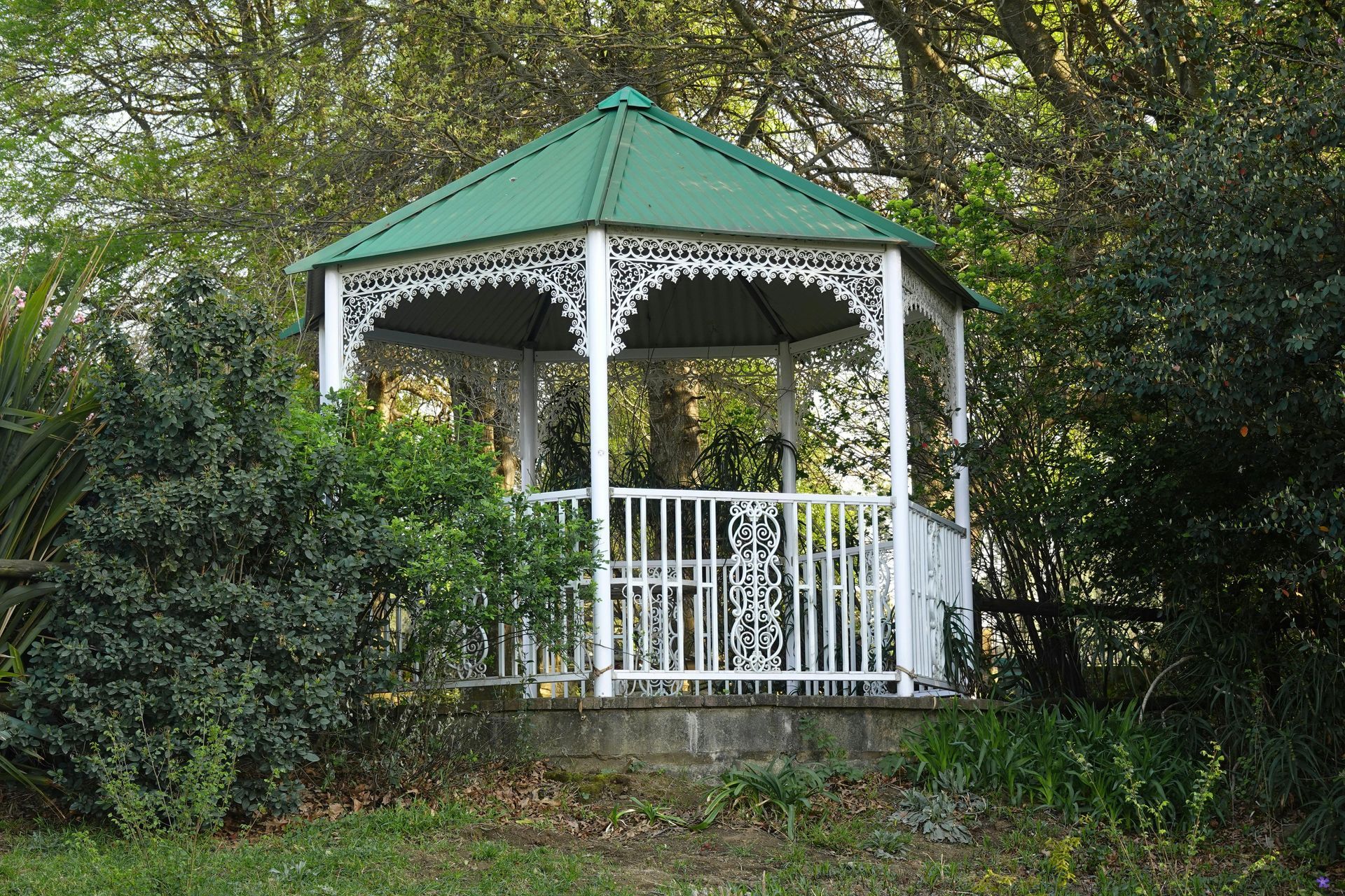 White gazebo with green roof in a garden, surrounded by bushes and trees.