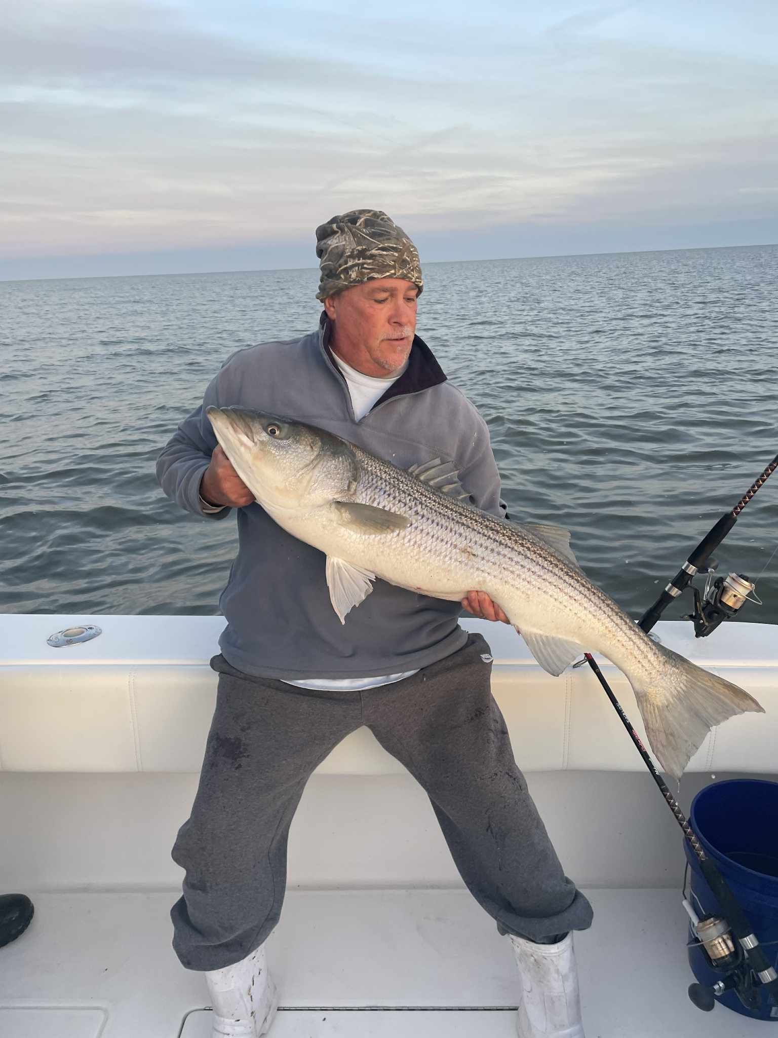 Man holding large striped bass on a boat, overcast sky, fishing rod present.