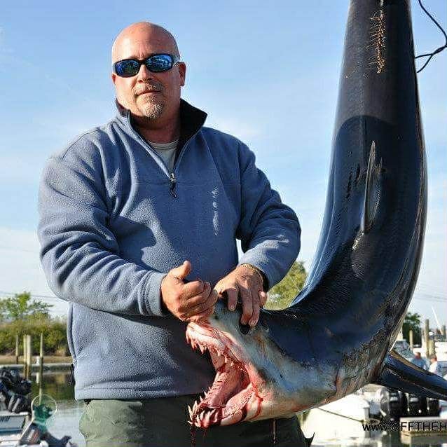 Man in sunglasses holds a large swordfish, mouth open, outdoors near boats.
