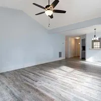 An empty room with pale blue walls, grey wood-look flooring, a ceiling fan, and an open doorway leading to a hallway.