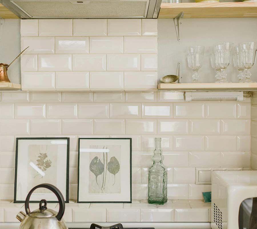 A modern kitchen featuring off-white subway tile walls, a tea kettle, framed botanical prints, and glassware on shelves.