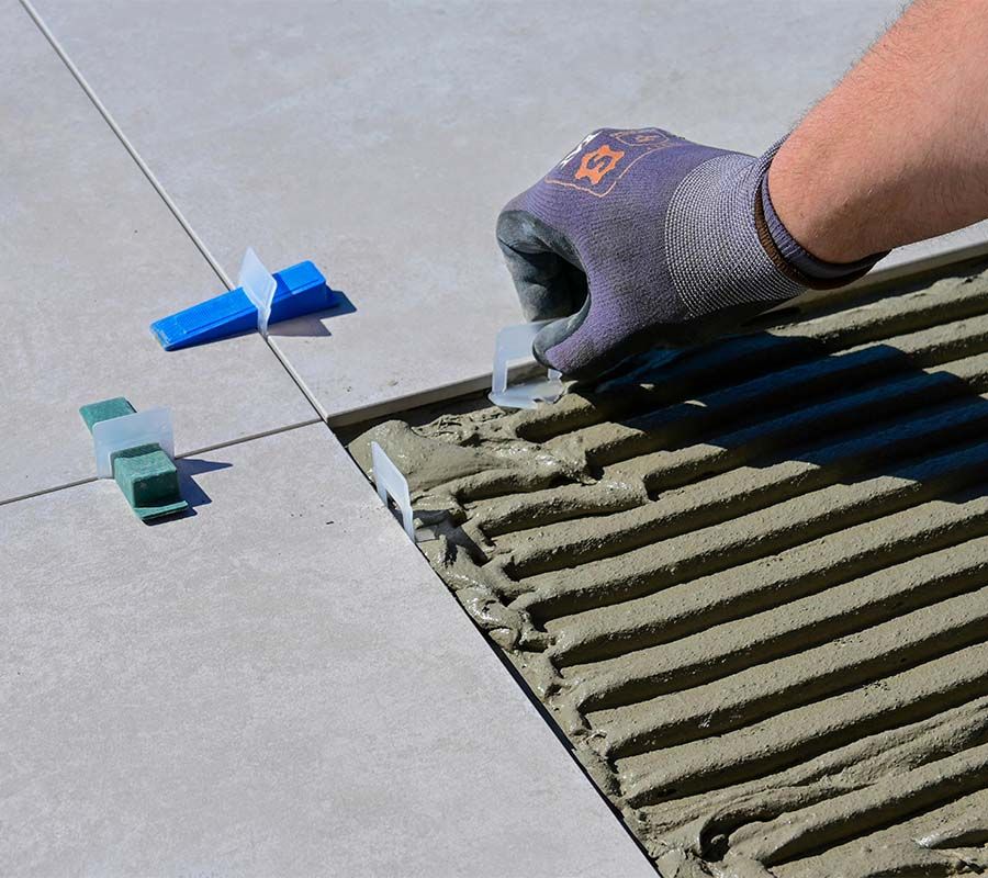 A gloved hand places a plastic tile leveling clip into wet mortar between two gray tiles on a construction site.