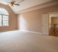 Empty bedroom with beige carpet and walls, vaulted ceiling, arched window, and open doorway leading to a wooden hallway.