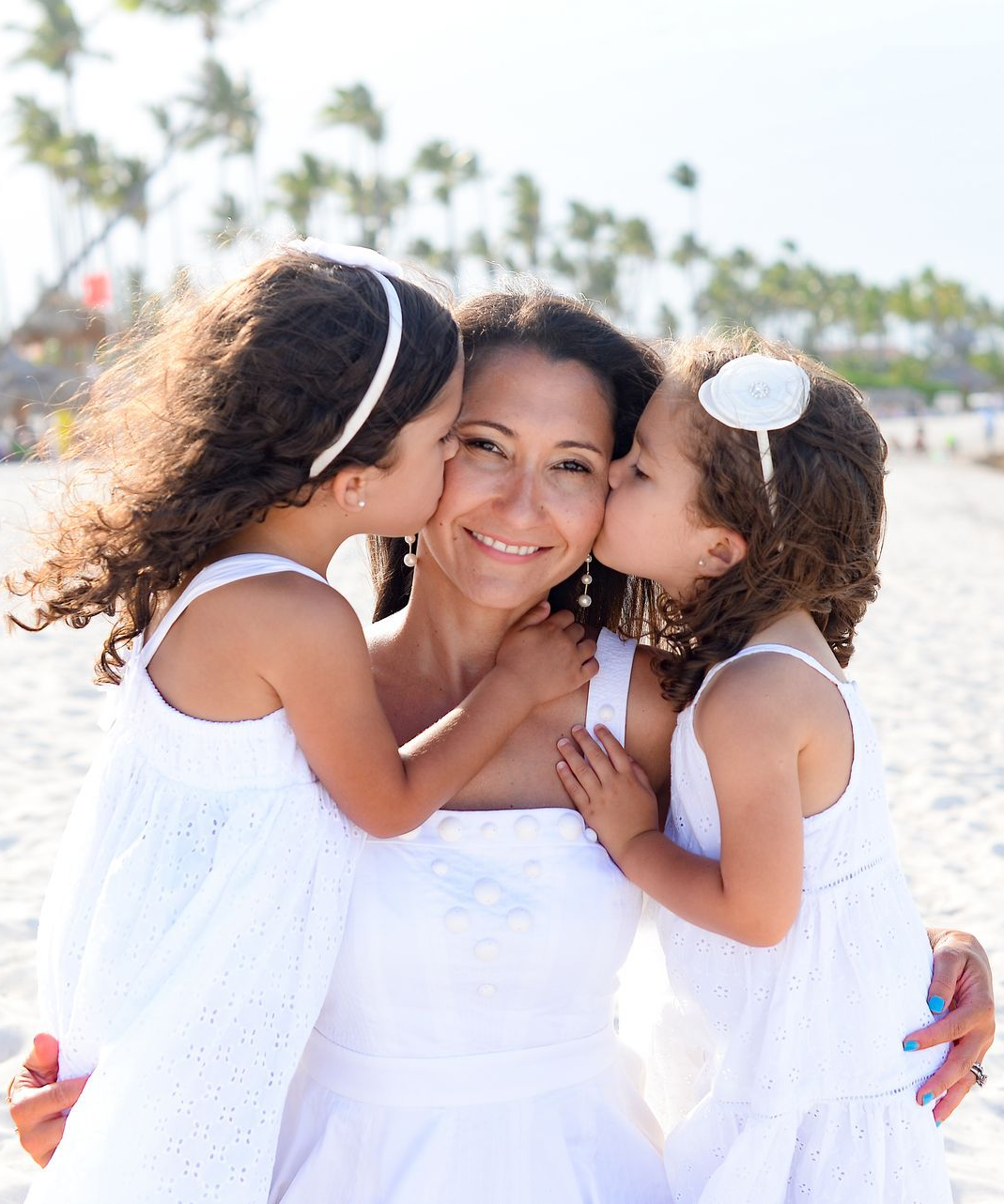 Mother on beach is kissed by two daughters, all in white dresses.