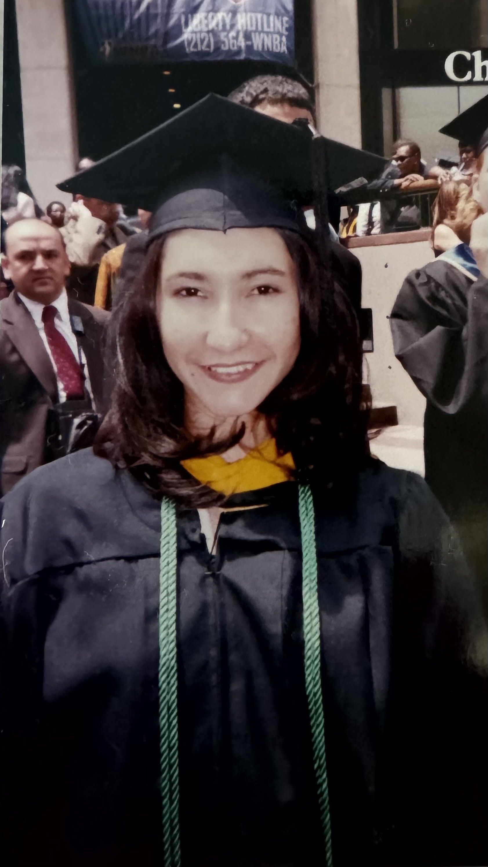 Woman in graduation cap and gown smiling, with a green and gold tassel, outdoors.