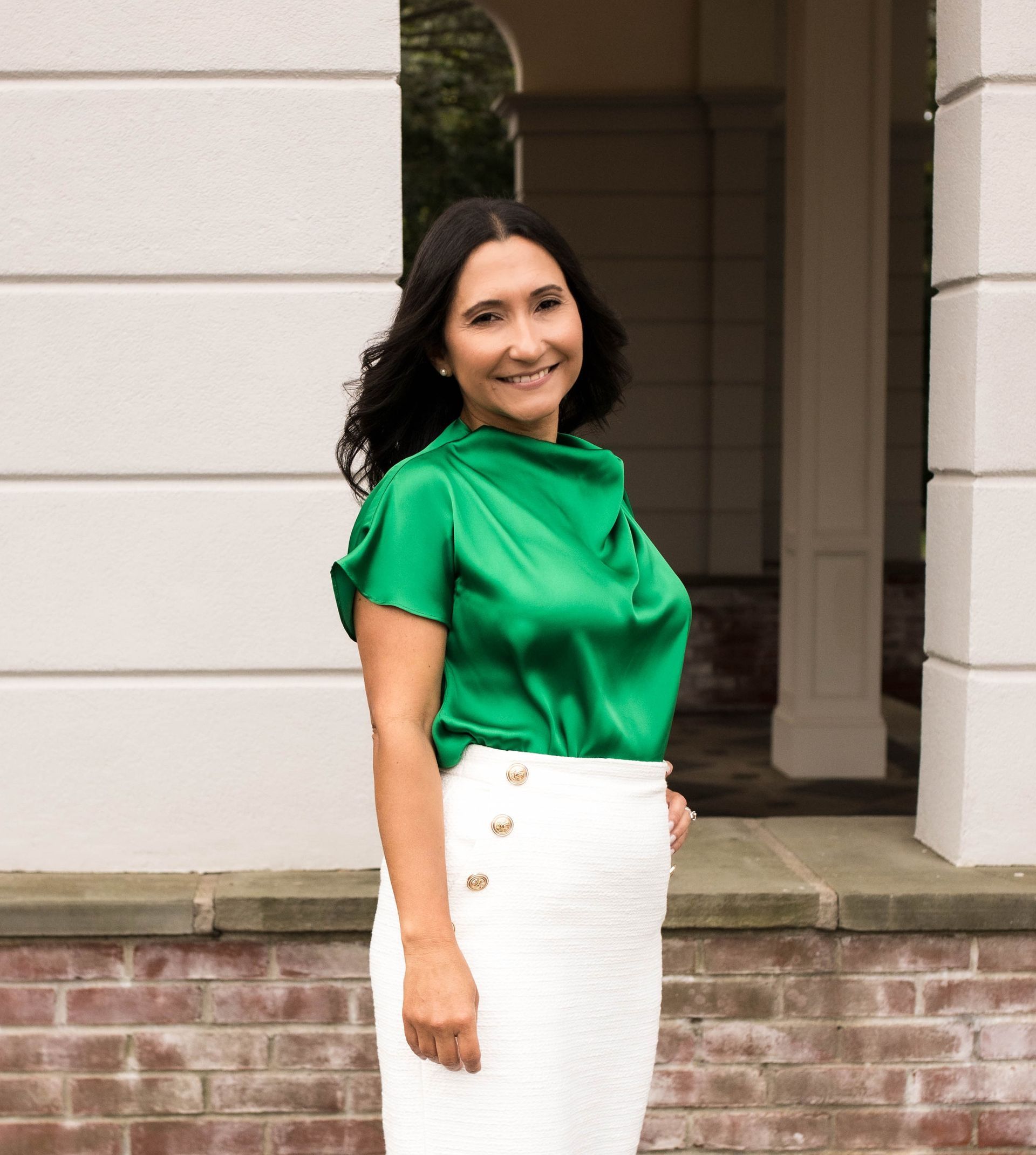 Woman in green top and white skirt smiles outdoors near a building.