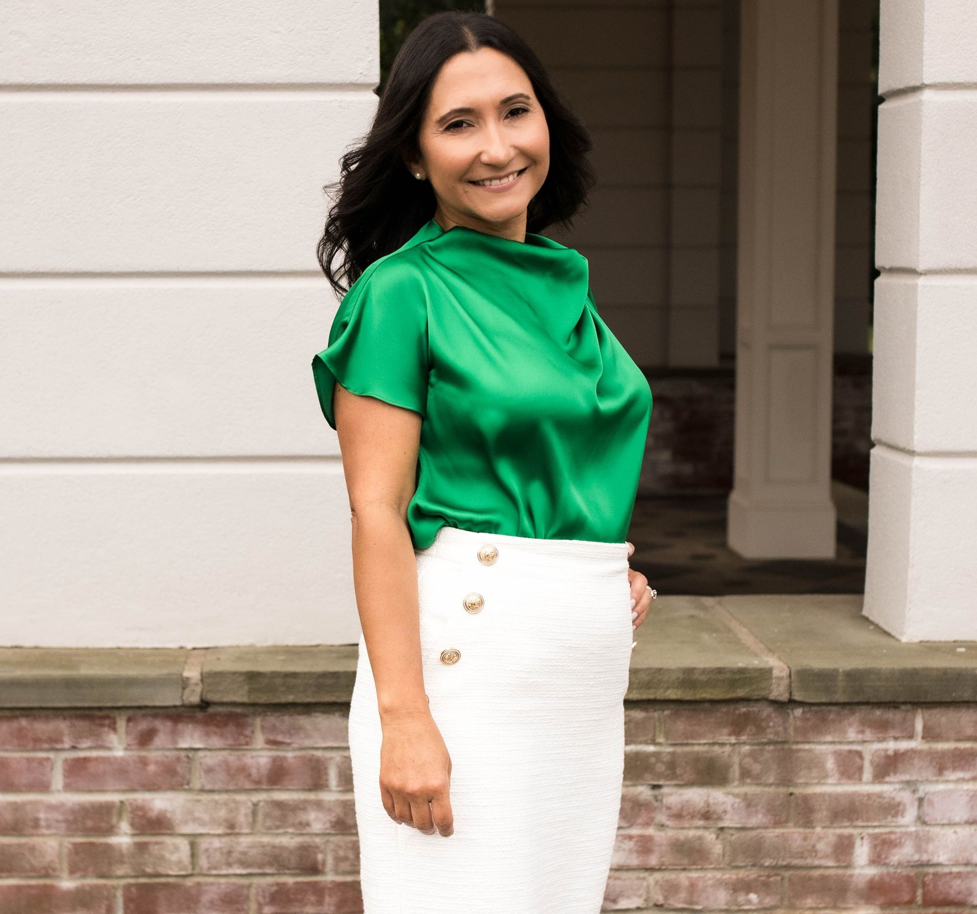 Woman in green top and white skirt smiles outdoors near a building.