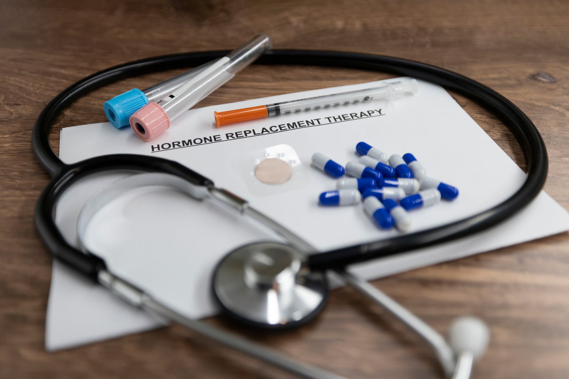 A stethoscope , syringe , pills , and a piece of paper on a wooden table.