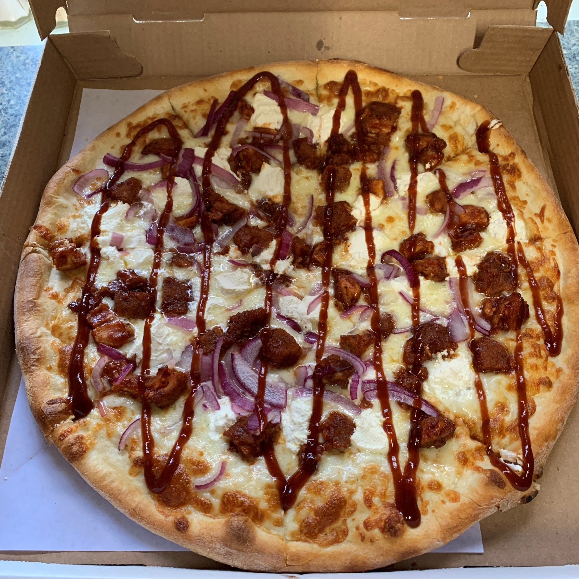 A pizza with meat , cheese , onions and barbecue sauce is in a box.