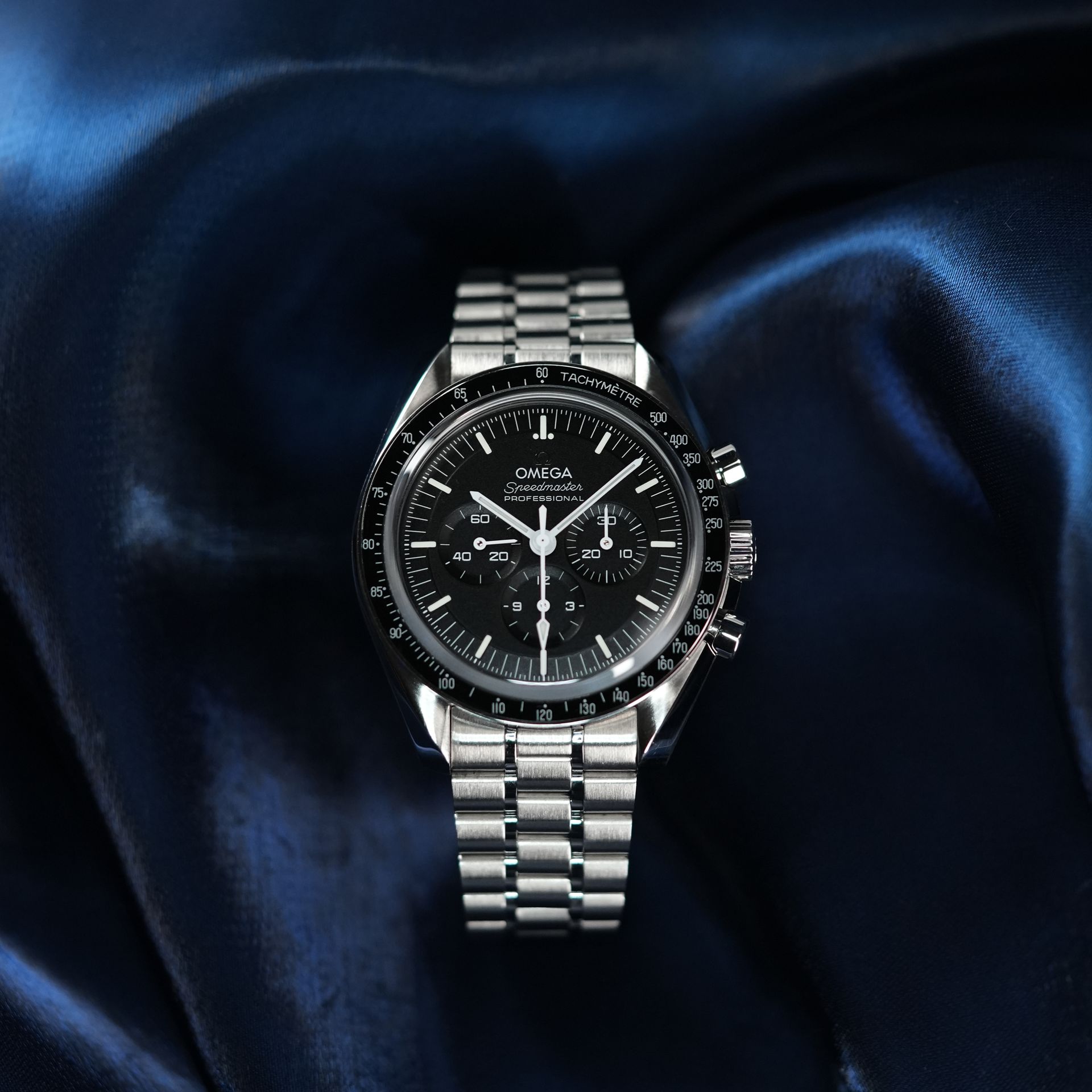 Omega Speedmaster watch with black face and silver band on a blue silk fabric.