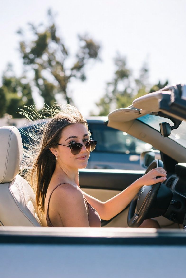 Eine Person mit langen braunen Haaren und Sonnenbrille lächelt, während sie an einem hellen, sonnigen Tag ein Cabrio fährt.