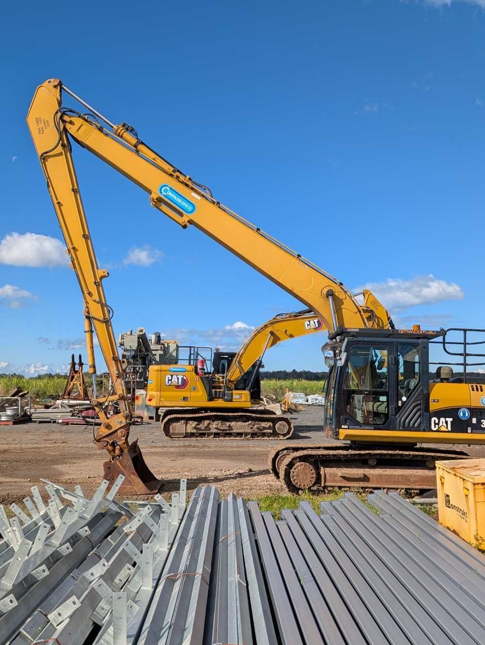 Yellow excavators working at a construction site with stacked metal beams under a blue sky — Compass Equipment Hire in Iluka, NSW