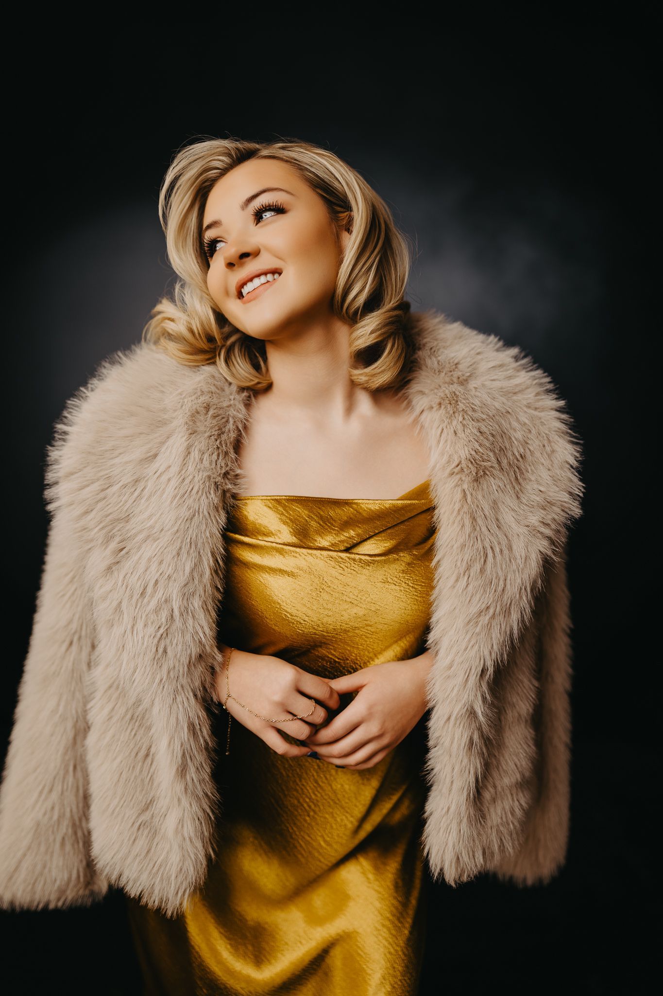A smiling person in a golden slip dress and a textured, beige faux-fur coat against a dark, moody background.