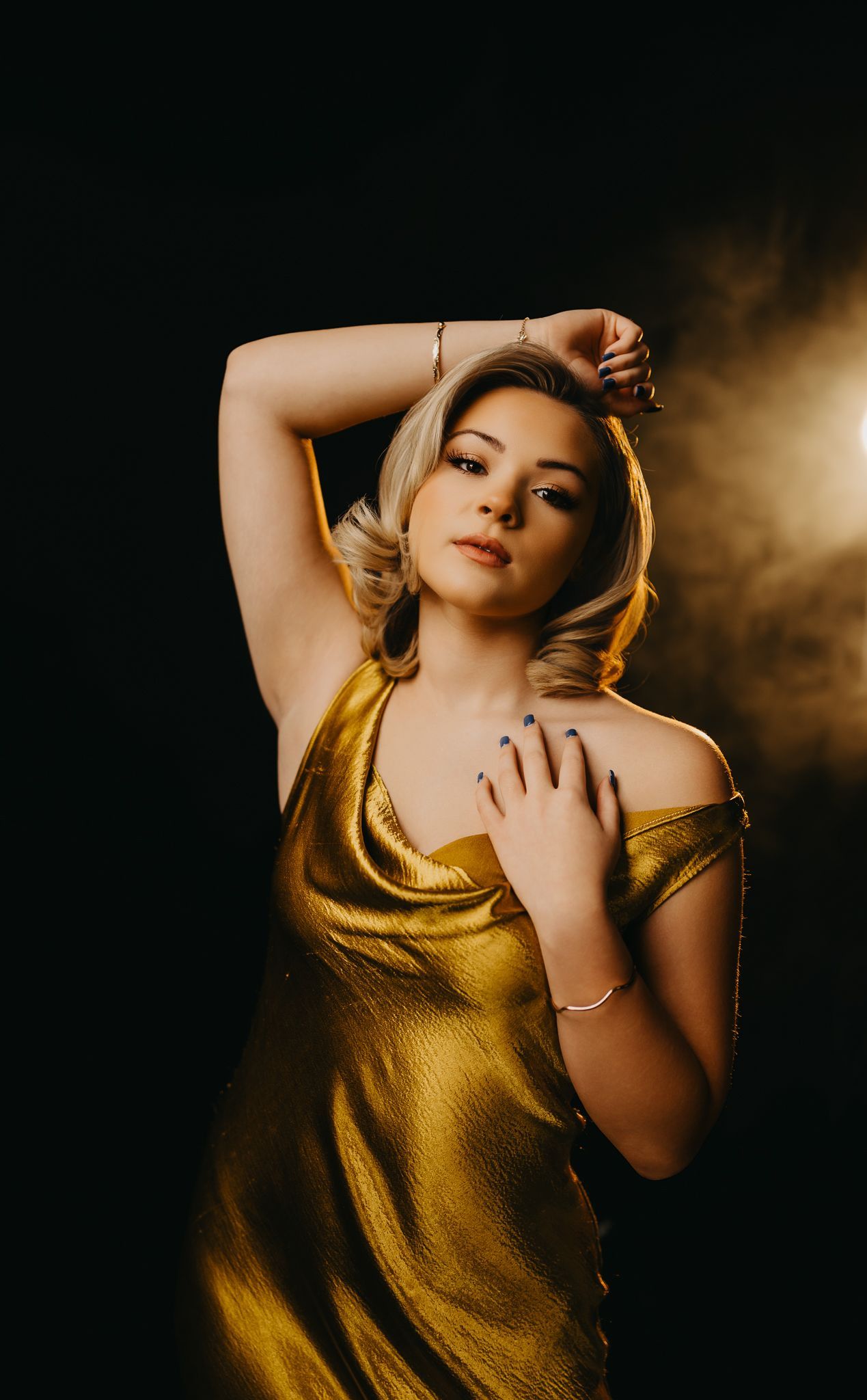 A person with light-colored hair poses against a black background, wearing a shimmering gold dress under soft lighting.