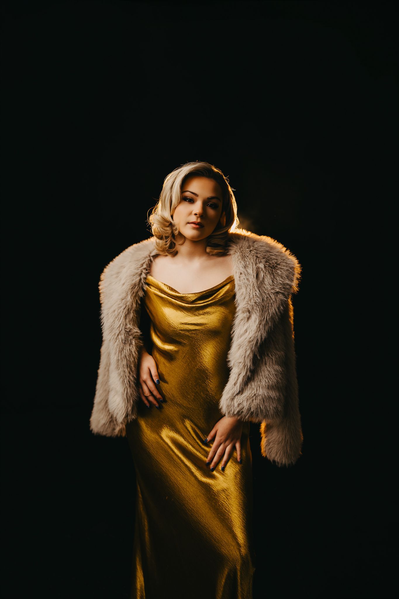 A person with wavy hair wears a gold satin slip dress and a shaggy faux fur coat against a solid black background.
