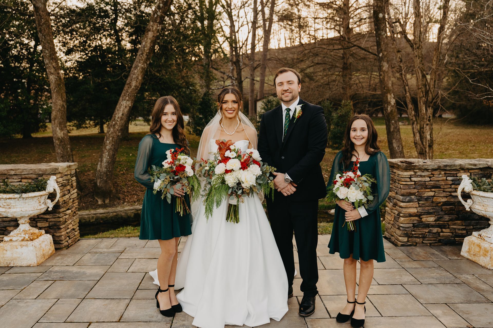 Bride and groom pose with two people in green dresses outdoors on a stone patio with large bouquets.