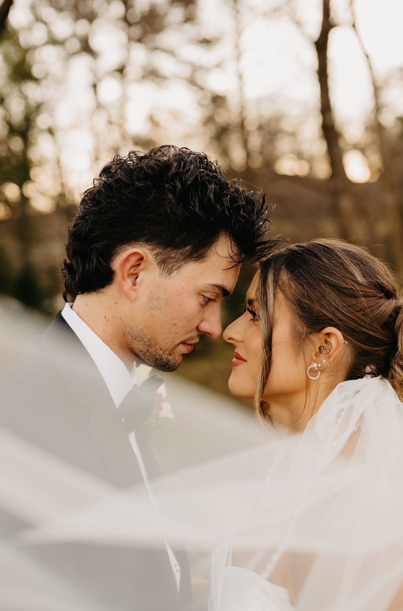 A couple gazes lovingly at each other during a wedding, partially obscured by a soft, flowing white veil in a forest.