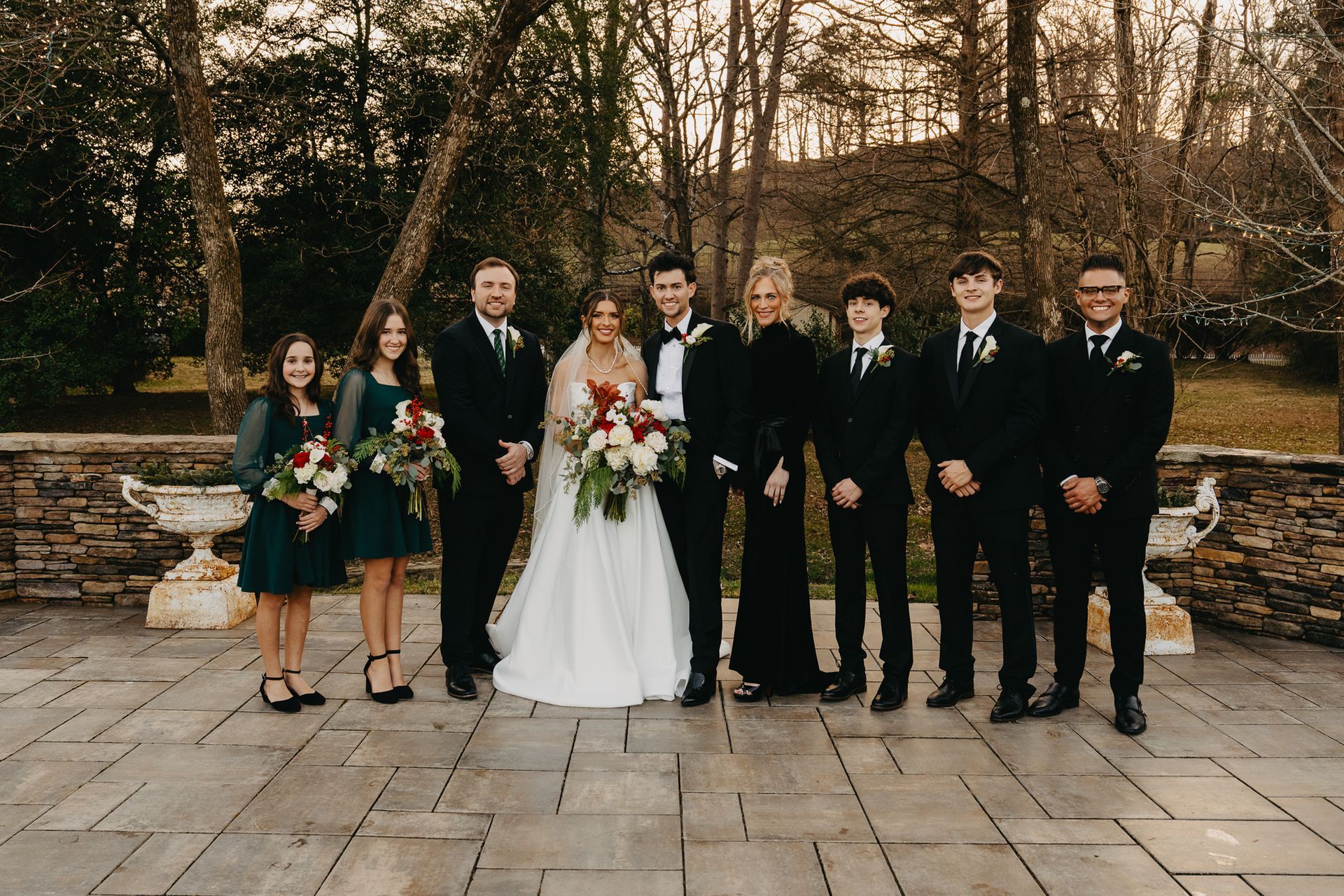 A wedding party stands outside on a stone patio in front of trees, including a bride and groom and six attendants.