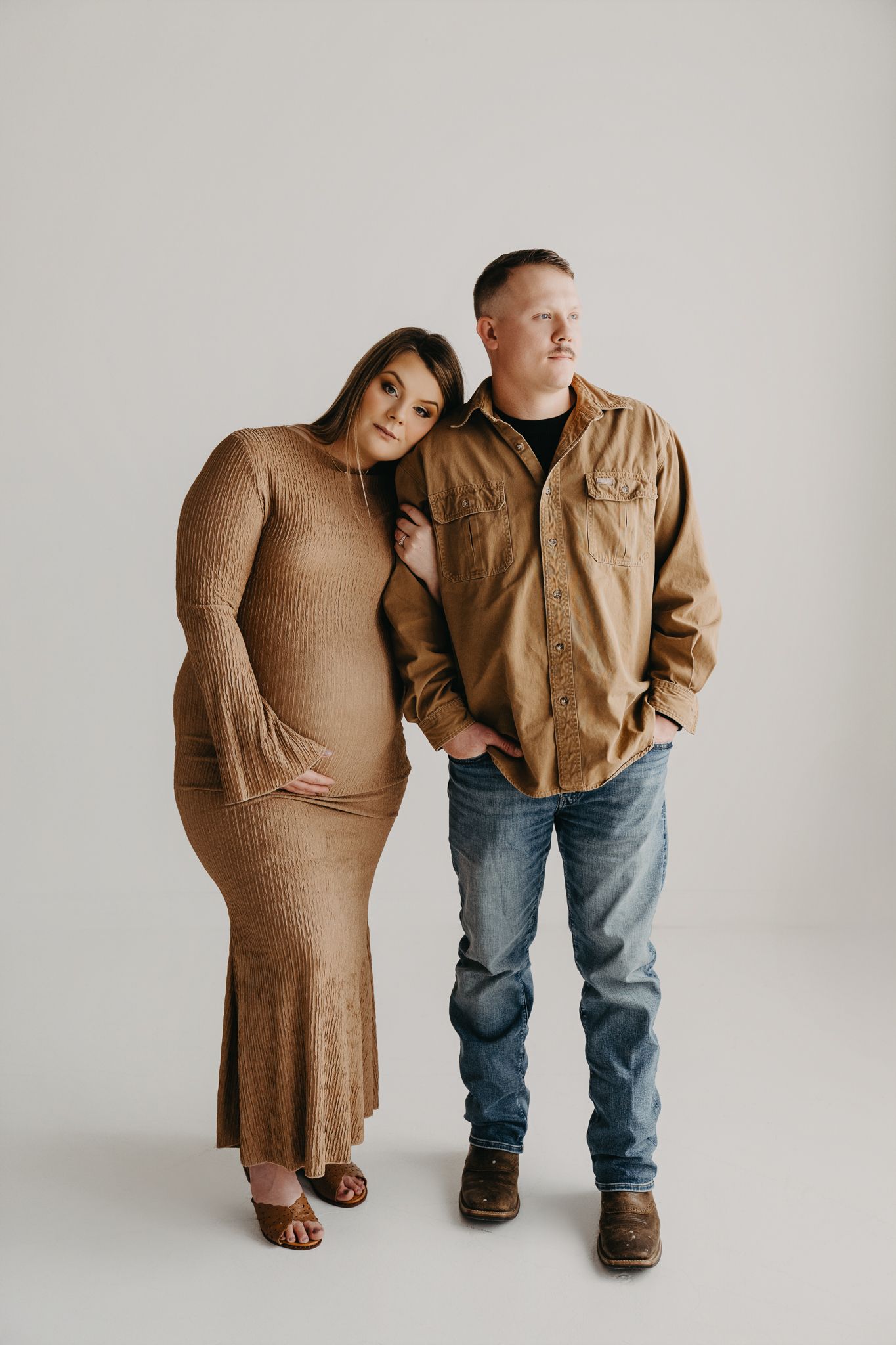 A person in a textured tan dress leans their head on the shoulder of a person wearing a tan shirt and blue jeans.