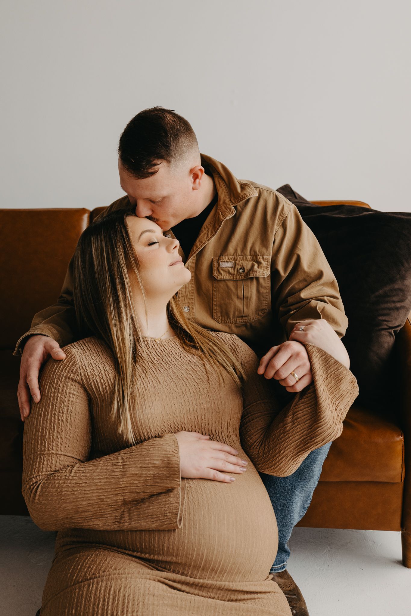 A couple embracing on a brown leather sofa, with one person resting a hand on the other's pregnant stomach.