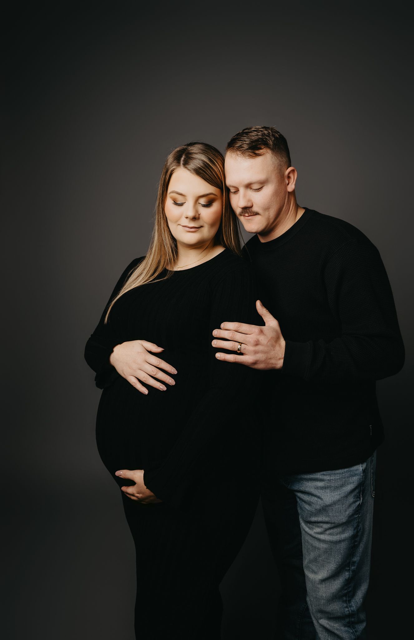 A pregnant person in a black long-sleeved dress and their partner in a black sweater standing against a dark backdrop.