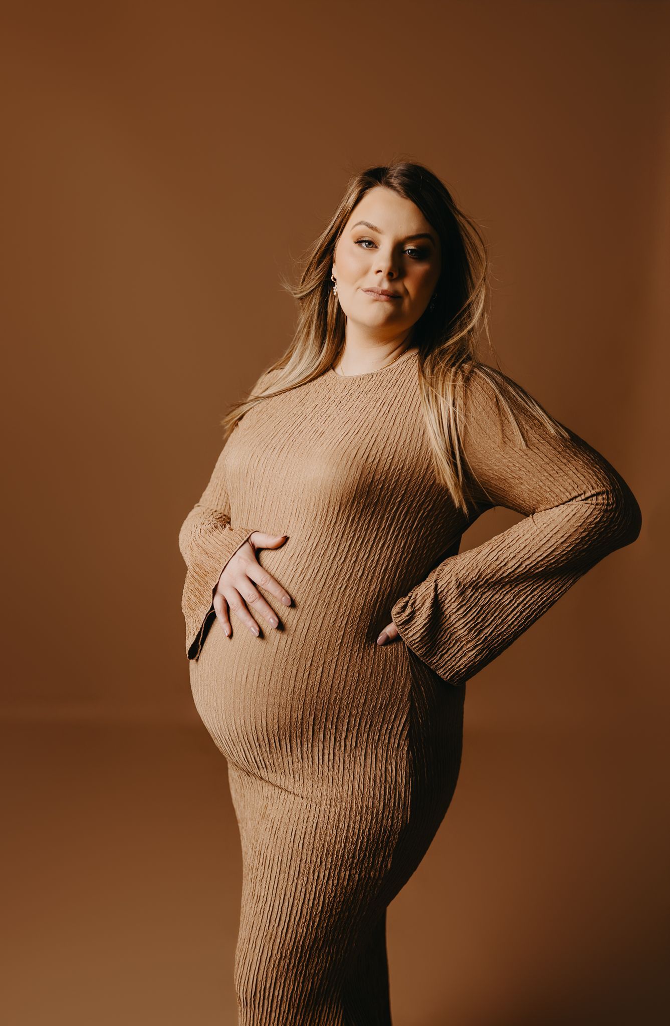 A person with long blonde hair poses against a solid tan background, wearing a textured, long-sleeved tan maternity dress.