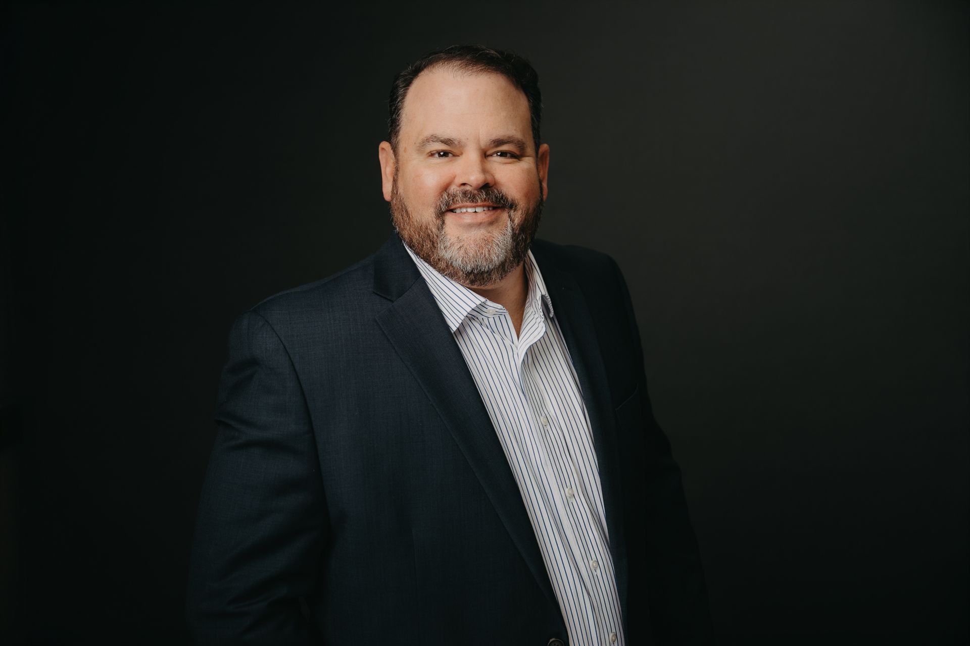 A smiling person in a dark suit and a patterned button-down shirt stands against a dark gray background.
