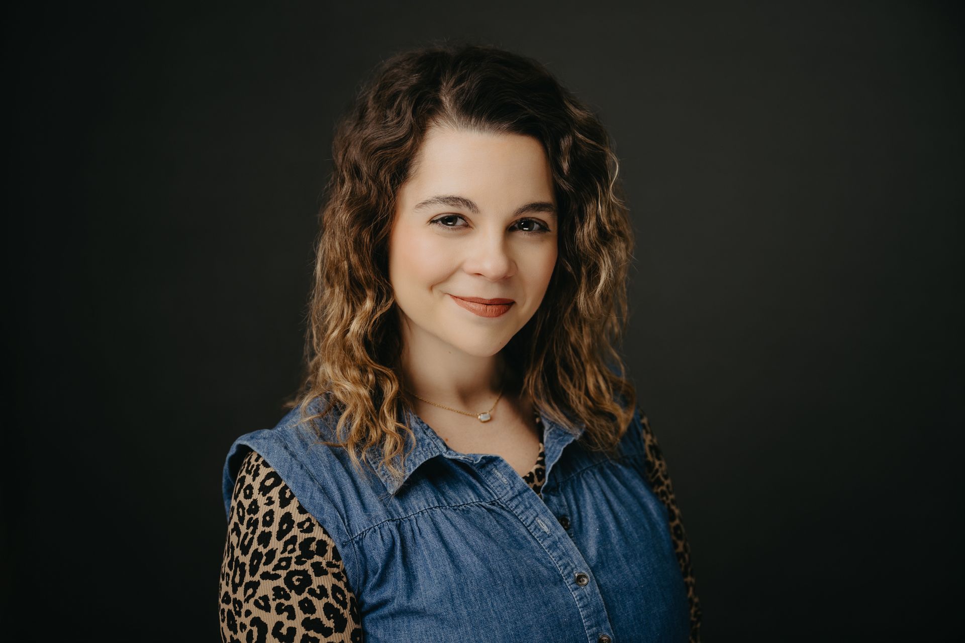 A person with shoulder-length curly hair wearing a denim vest over a leopard print shirt, smiling against a dark background.