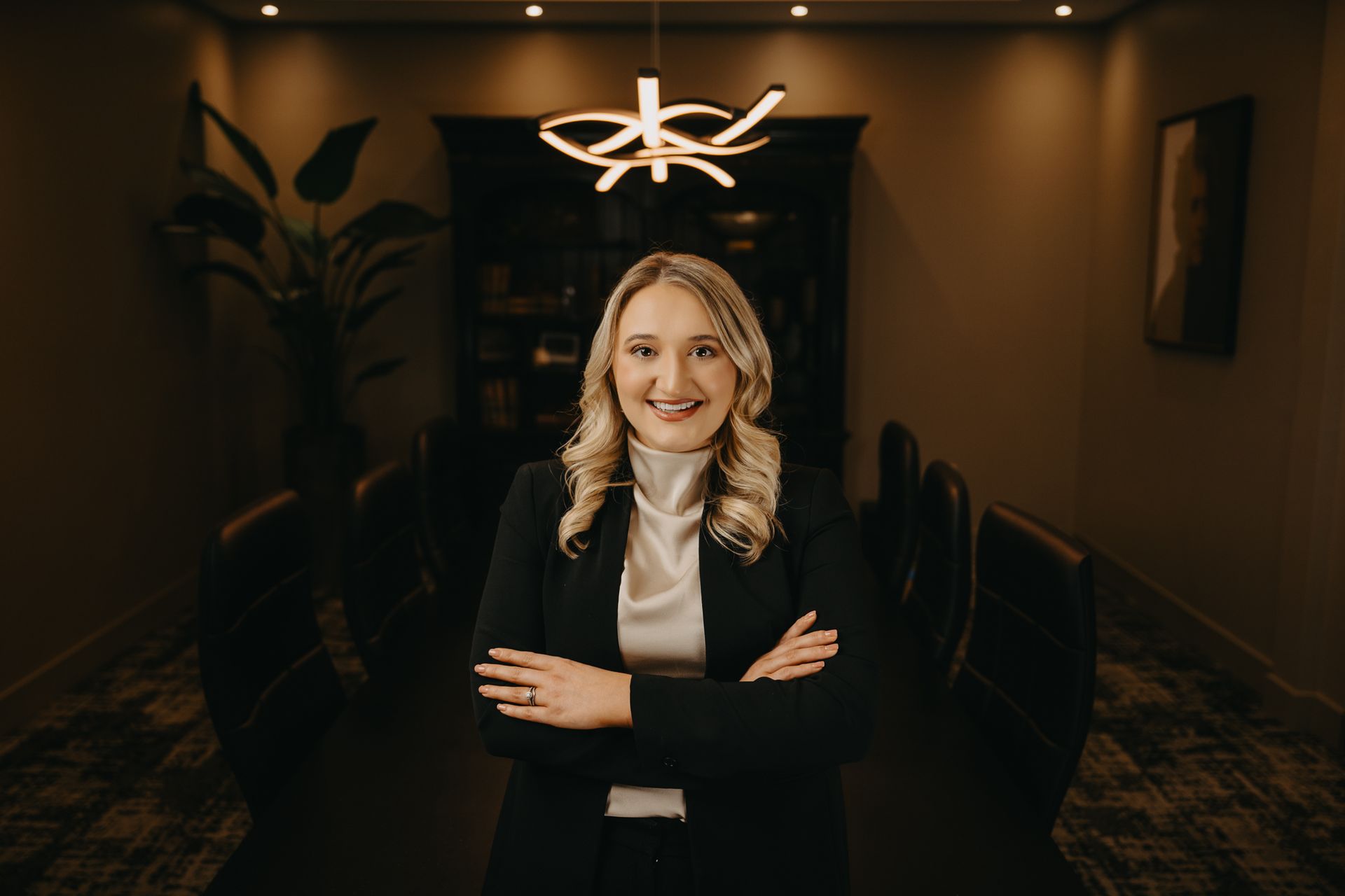 A professional with arms crossed, wearing a blazer and turtleneck, stands in a dimly lit boardroom with a modern light.