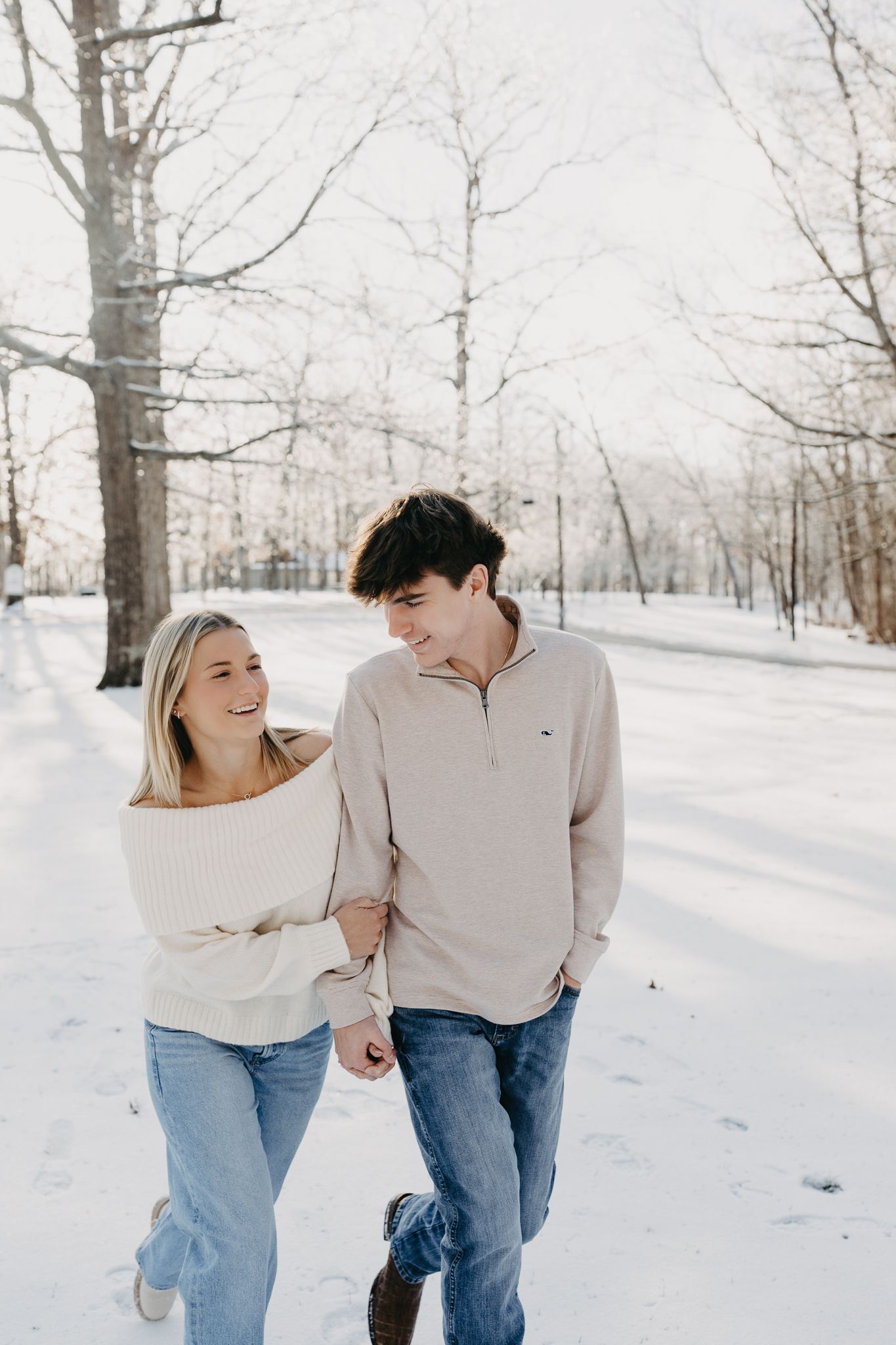 A young couple walks arm-in-arm through a snow-covered park, smiling at each other.