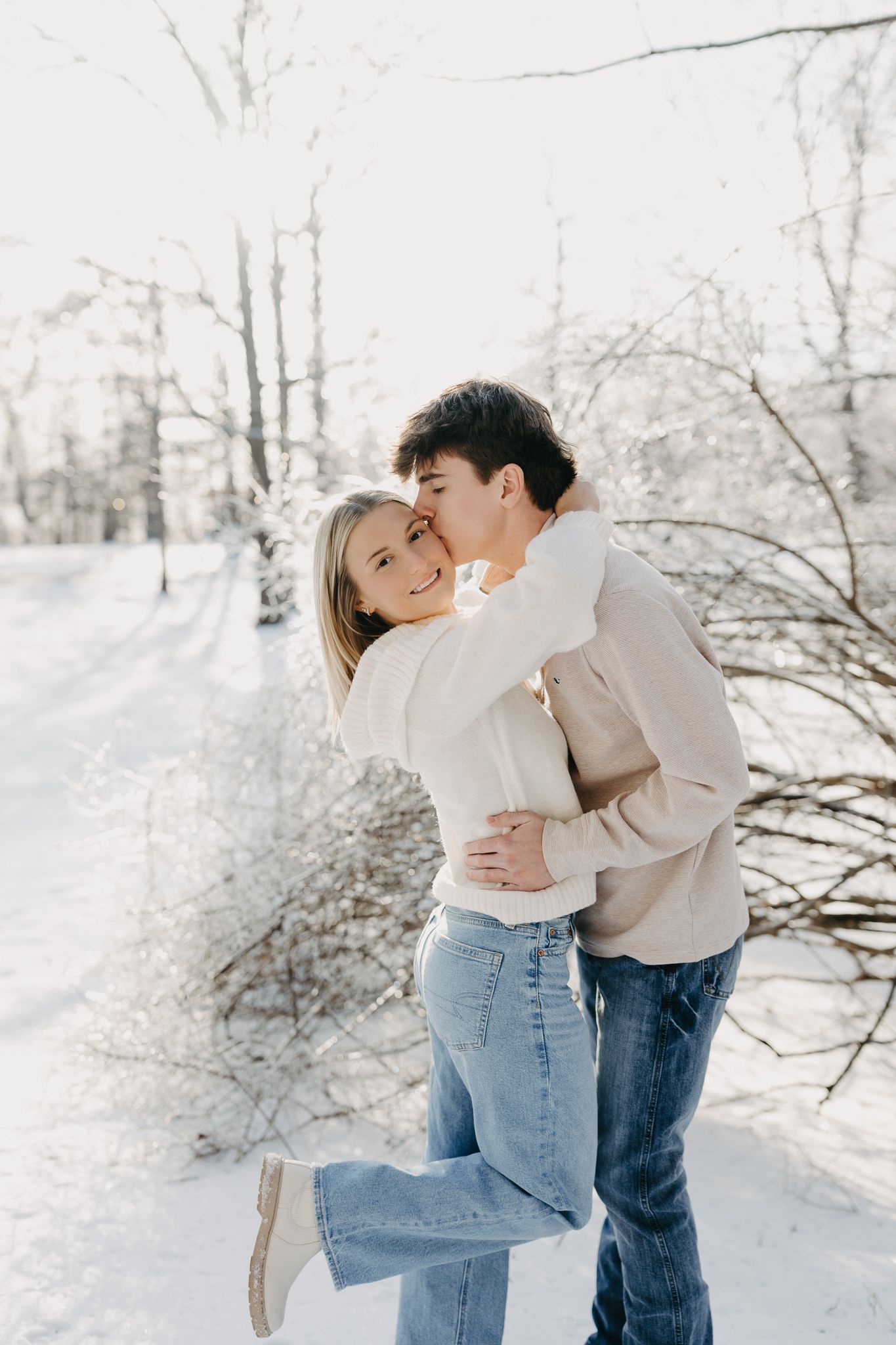 A couple wearing sweaters and jeans embraces in a snowy outdoor setting as one kisses the other on the cheek.