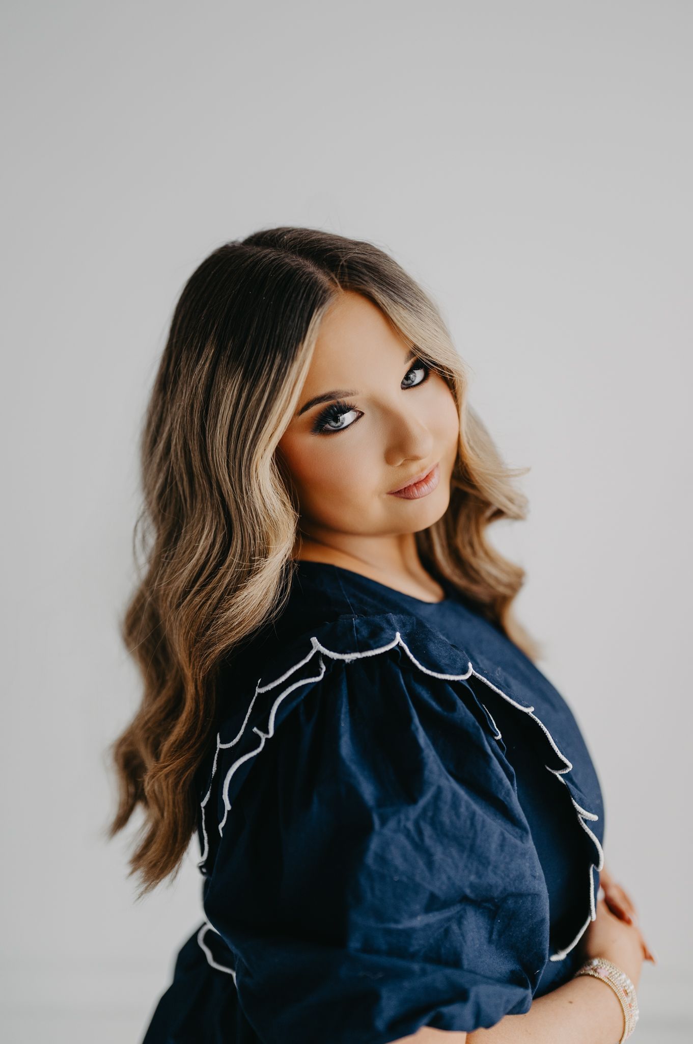 A person with long wavy hair and heavy makeup wearing a dark blue blouse with white trim, posed against a gray backdrop.