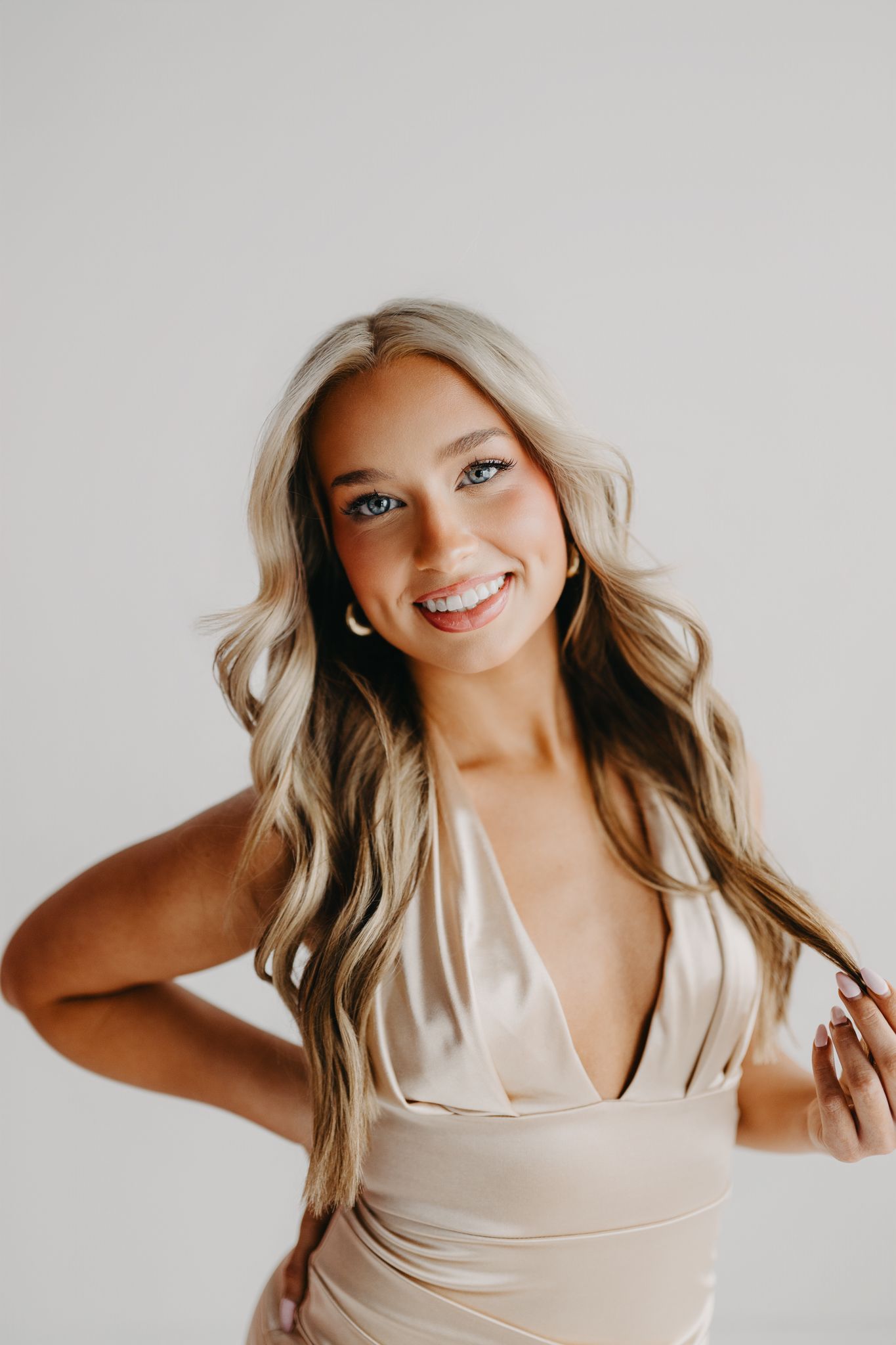 A smiling person with long blonde hair, wearing a light champagne-colored dress, poses against a plain white background.