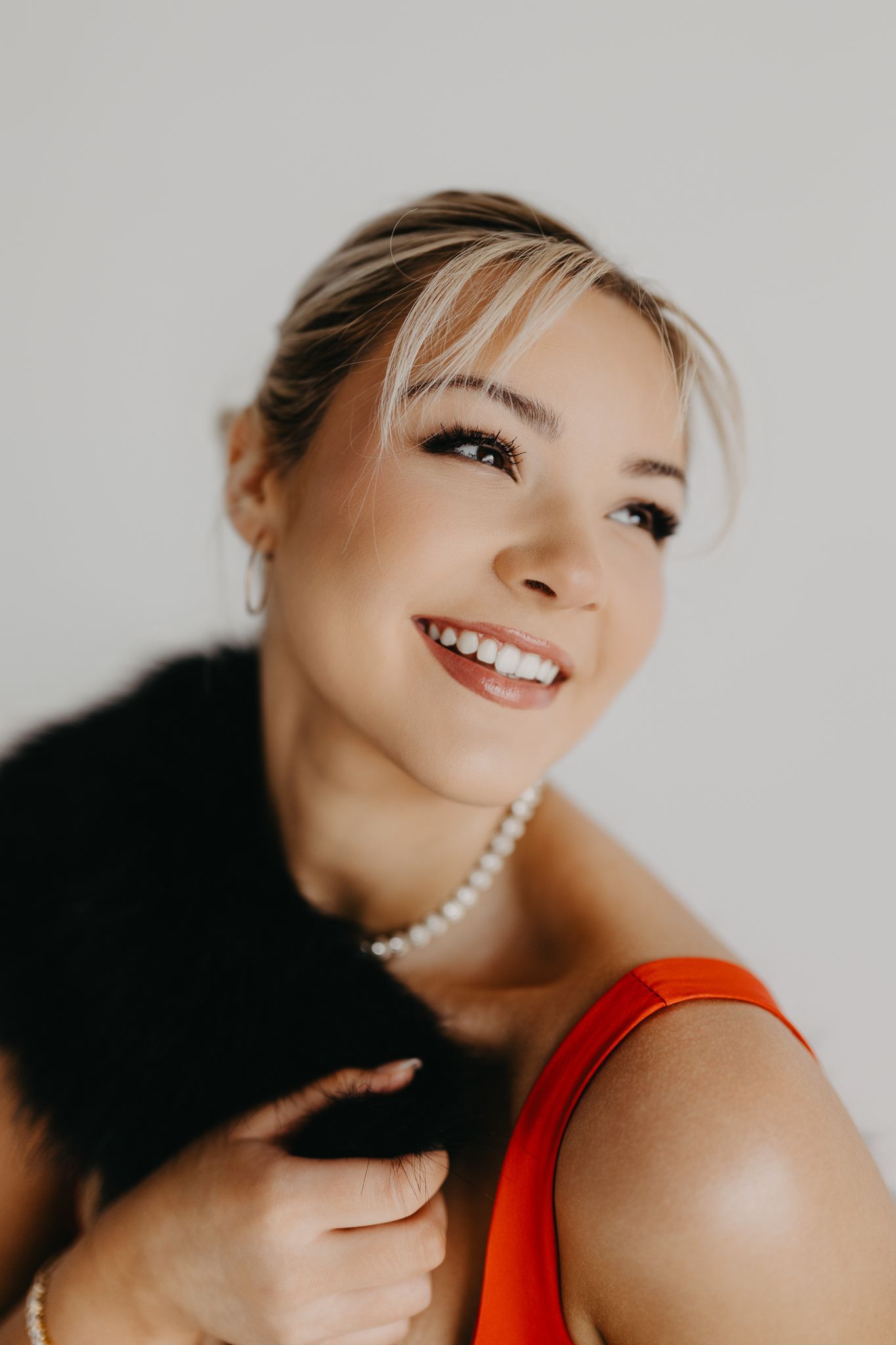 Smiling person wearing a red top, pearl necklace, and a black faux fur stole against a plain white background.