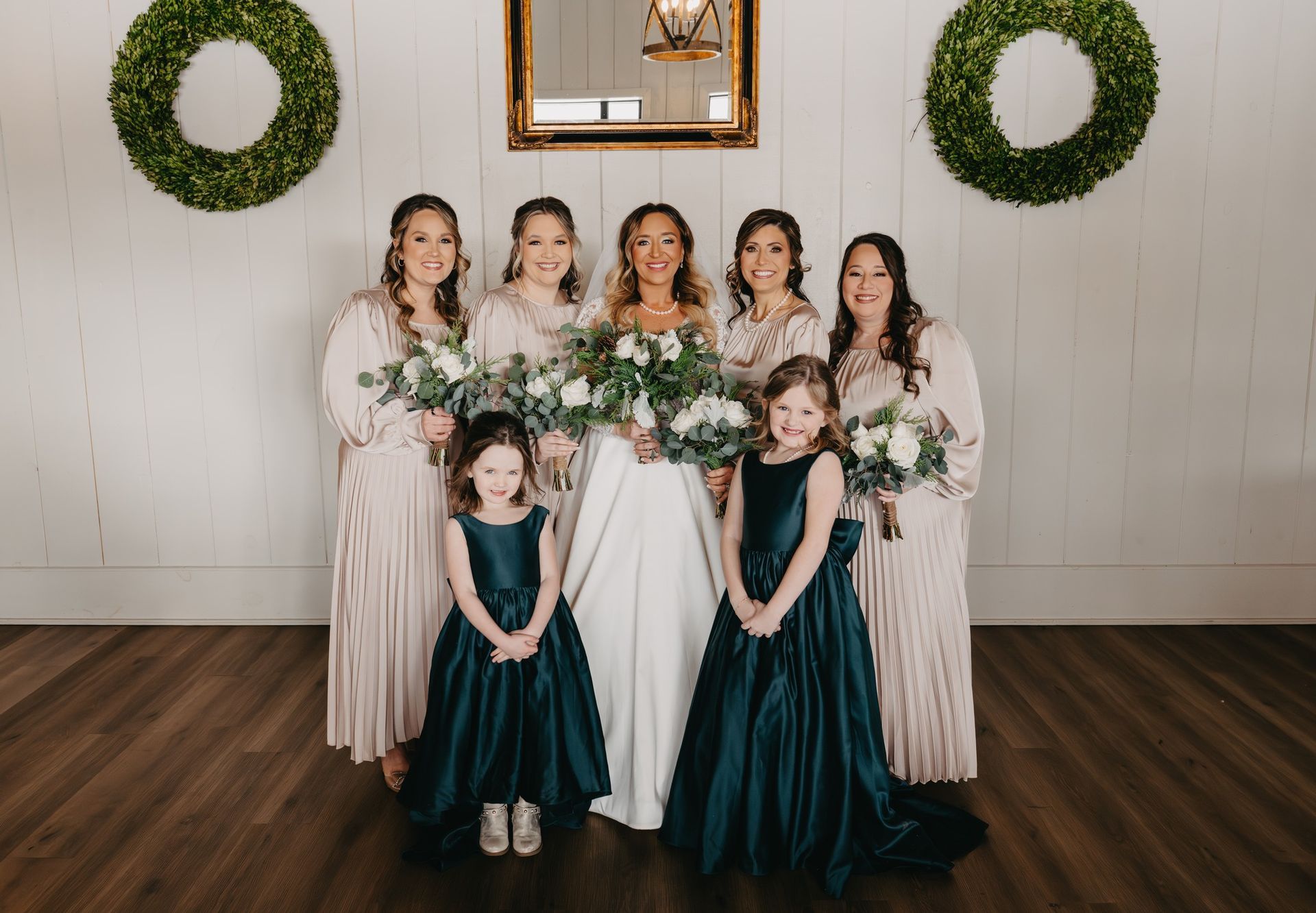 A bridal party poses against a white wall with greenery wreaths; bridesmaids in rose dresses and two girls in teal.