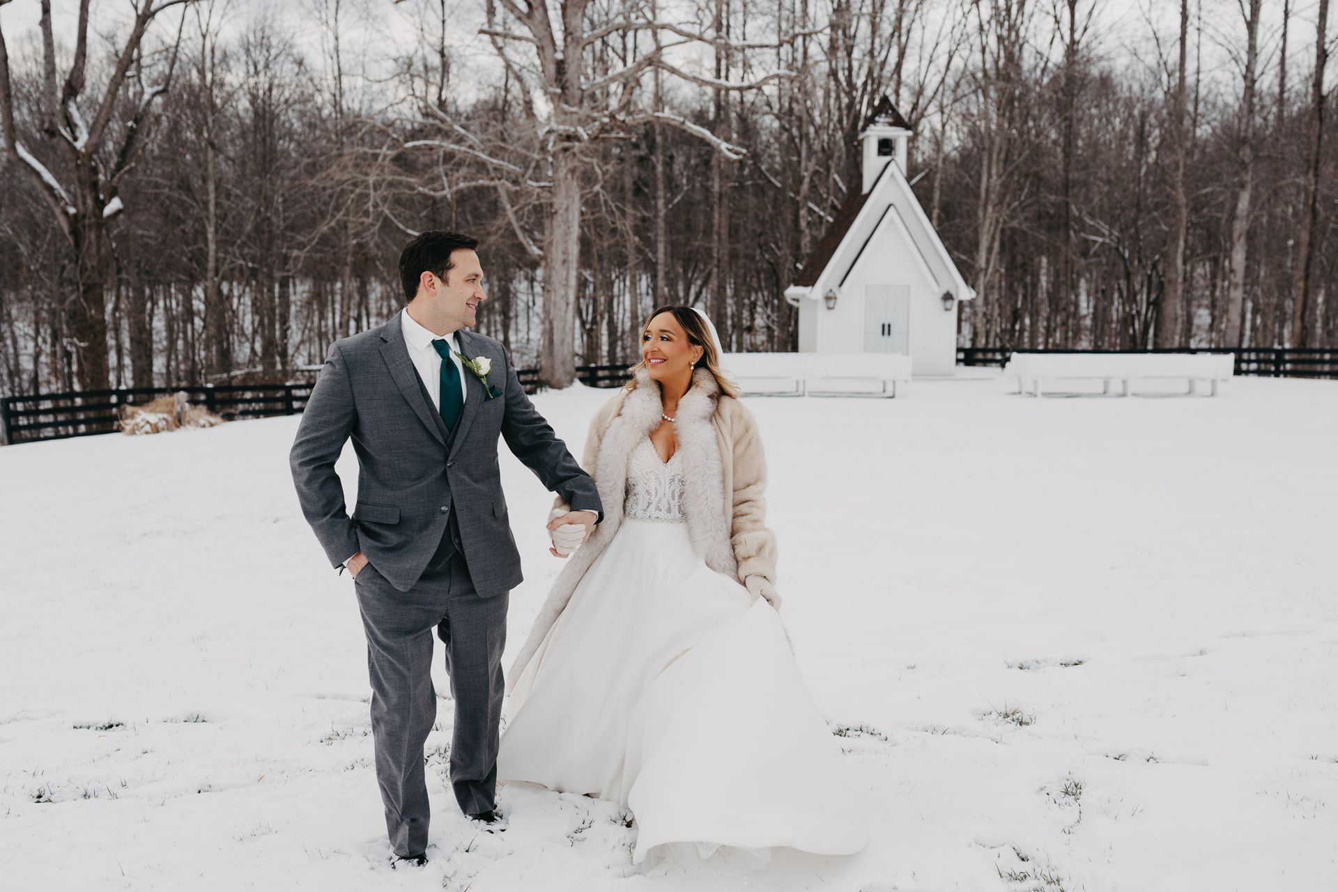 A couple in wedding attire walks hand-in-hand across a snow-covered field toward a small white chapel.