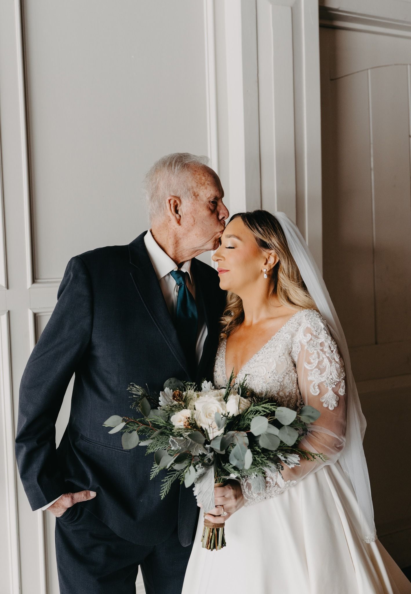 A bride in a white lace-sleeved gown holding a bouquet stands while someone kisses her forehead in a bright room.