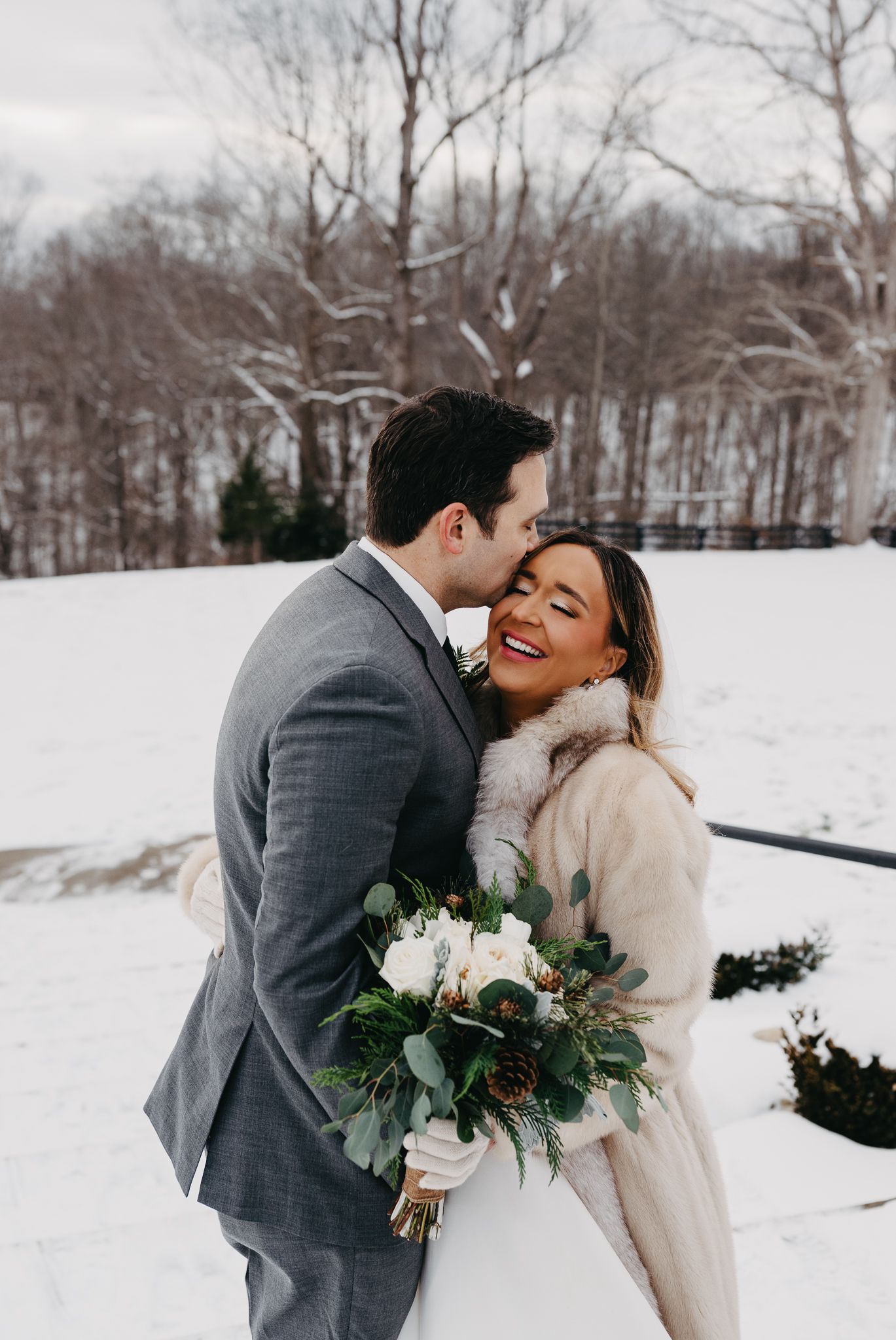 A person in a dark suit kisses the forehead of a smiling person in a cream coat holding a bouquet in a snowy field.