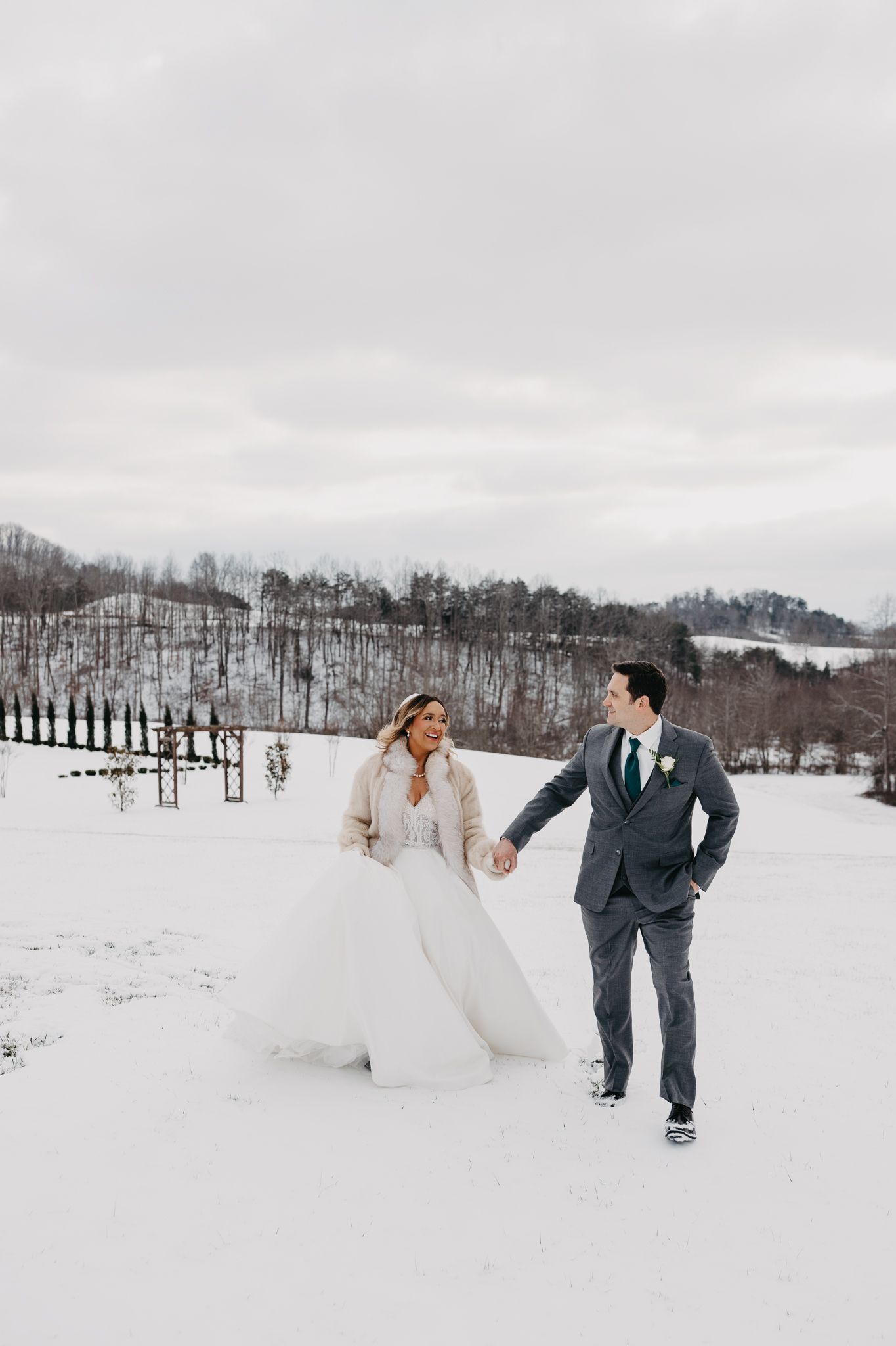 A couple in wedding attire walks hand-in-hand through a snow-covered field under a cloudy sky.